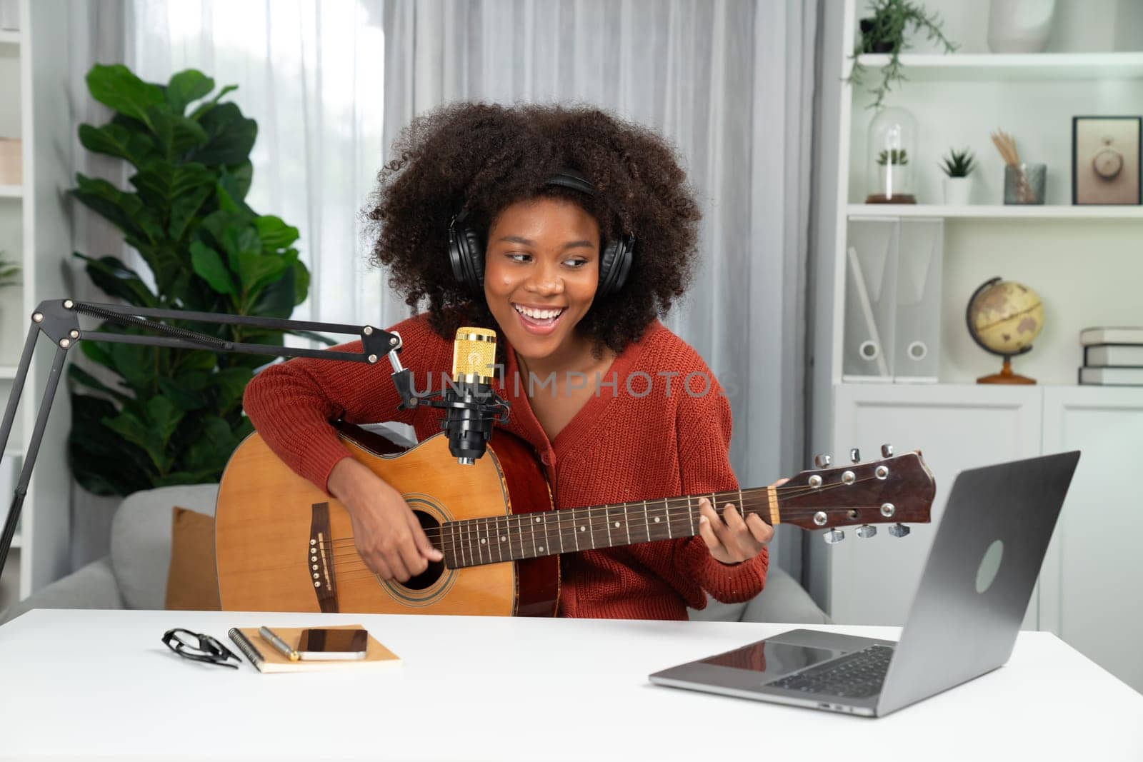 Host channel in musician of young African American playing guitar along with singing, broadcasting on laptop in studio. Decoration of equipment of headsets and recording microphone. Tastemaker.