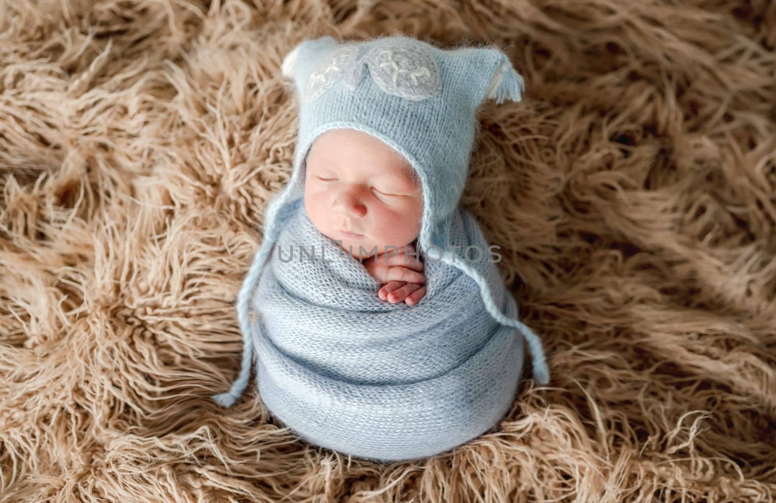 Adorable newborn baby boy swaddled in knitted blanket and wearing hat on fur studio portrait. Cute infant child kid sleeping