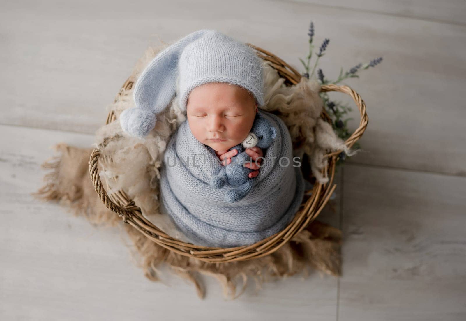 Adorable newborn baby boy swaddled in knitted blanket holding teddy bear toy and sleeping in basket studio portrait. Cute infant child kid napping