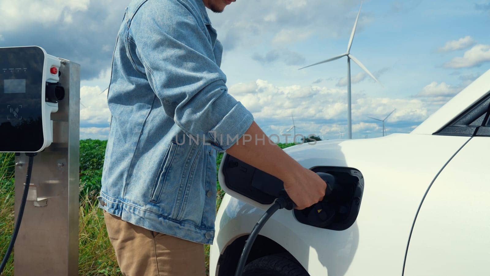 Progressive man with his electric car, EV car recharging energy from charging station on green field with wind turbine as concept of future sustainable energy. Electric vehicle with energy generator.