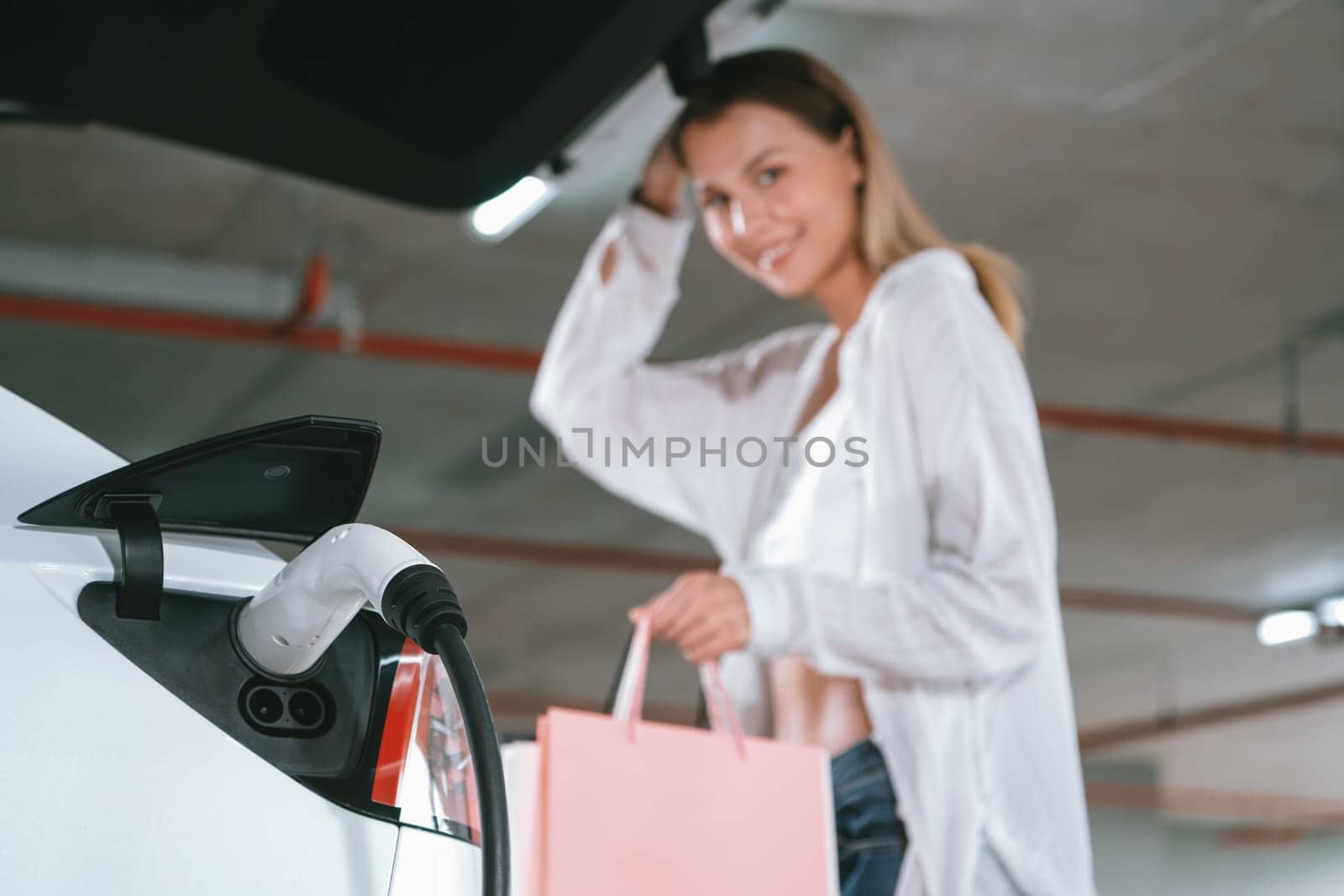 Young woman travel with EV electric car to shopping center parking lot charging in downtown city showing urban sustainability lifestyle by green clean rechargeable energy of electric vehicle innards