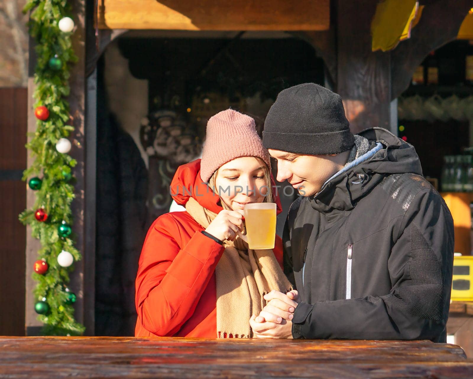 Happy student girl and boy drinking hot mulled wine and smiling at Christmas market. High quality photo. Photo of joyful young couple speending weekend together in winter. Portrat of two posivite amazed partners drinking a hot drink at the Christmas