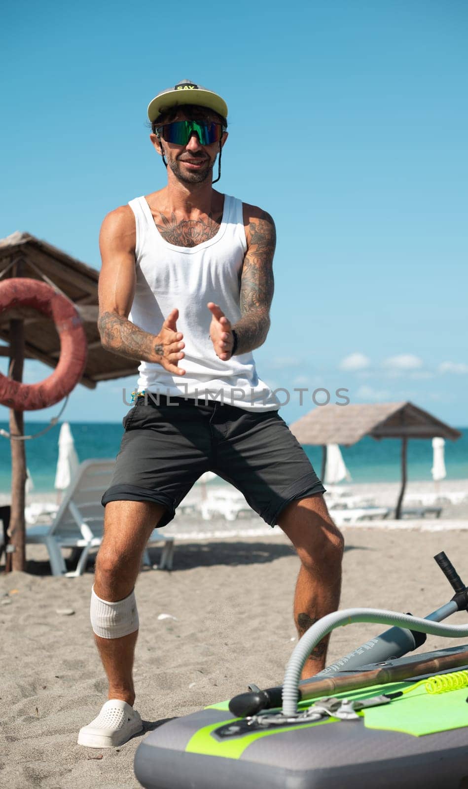 a sporty surfer guy with a pump pumps up an inflatable SUP board for swimming on the seashore by Rotozey