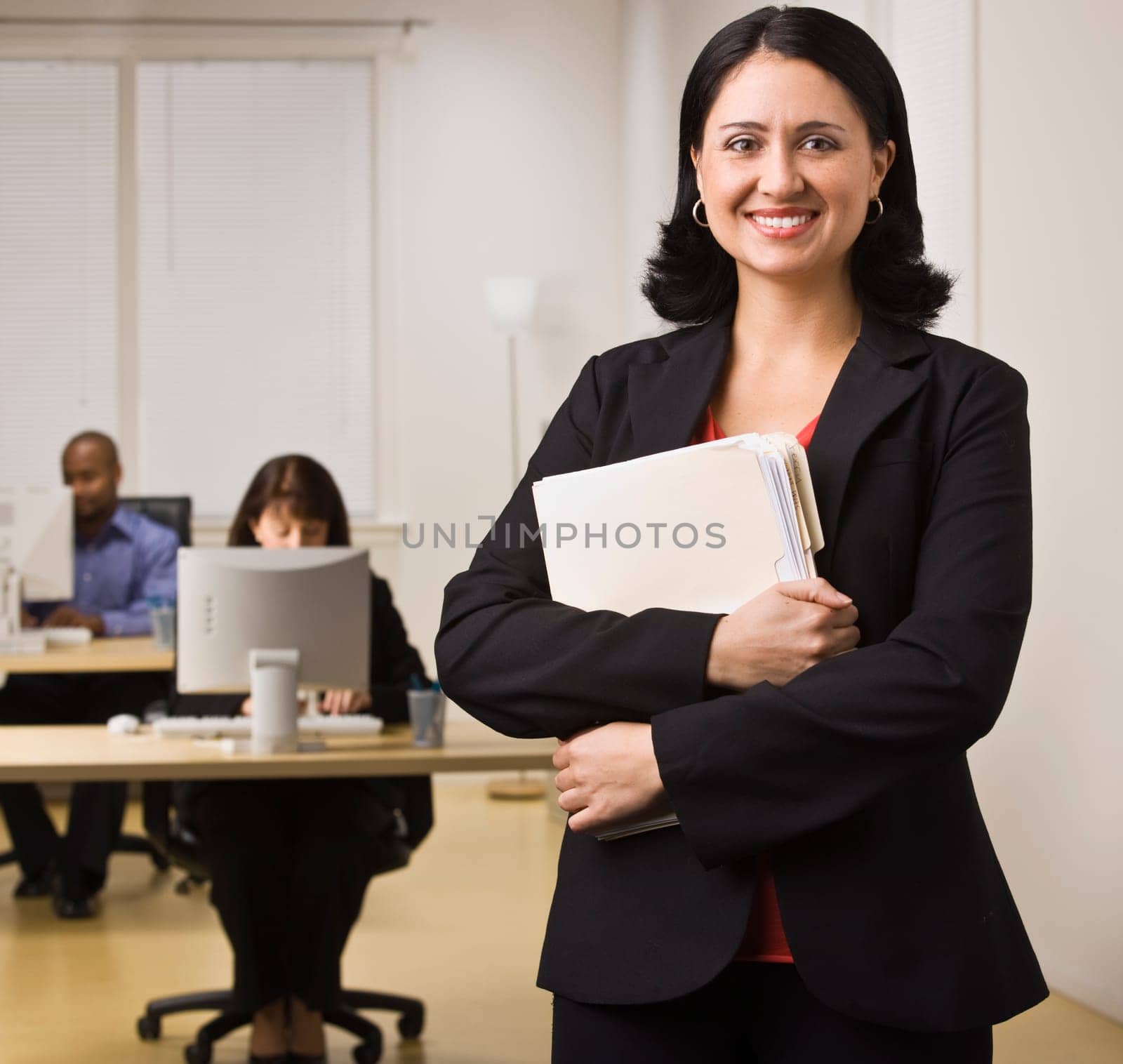 An attractive brunette is holding file folders and is smiling at the camera. She is dressed in business attire and appears to be at her place of employment. There are people working on computers at the desks behind her. Square composition.