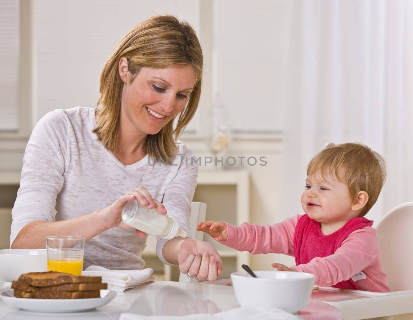 Smiling mother and baby daughter sit together at the breakfast table as the mother tests the temperature of the baby's bottle. Square framed shot.