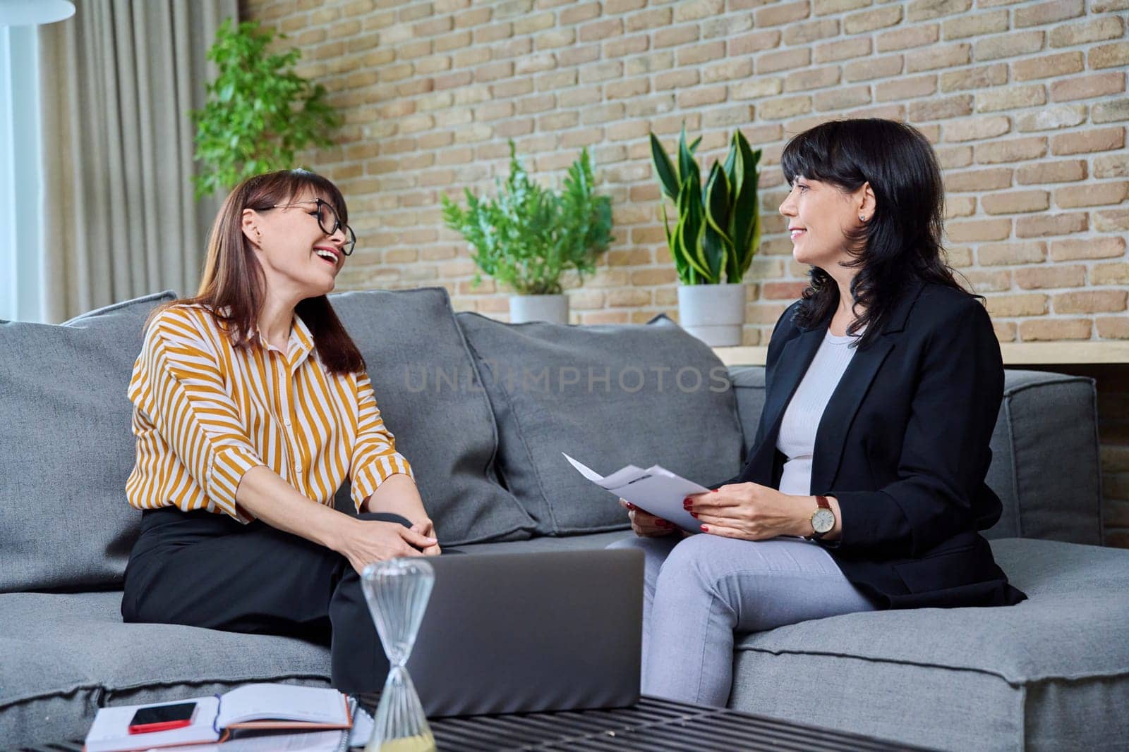 Two mature business women are talking in a modern office. Laughing communicating female colleague, 40s 50s age. Business, ceo, work, law, finance, mentoring, consulting, teamwork, job