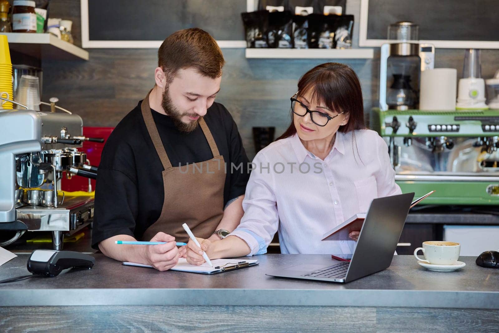 Team, partners, young male and mature woman talking working using laptop standing behind bar in coffee shop. Team, small business, work, staff, cafe cafeteria restaurant, entrepreneurship concept