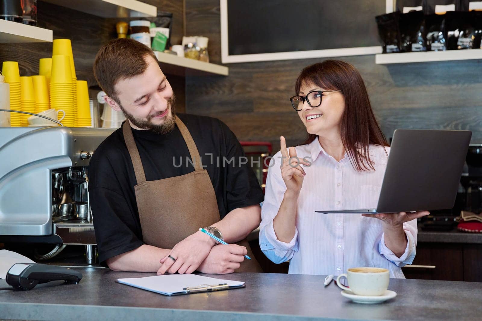 Team, partners, young male and mature woman talking working using laptop standing behind bar in coffee shop. Team, small business, work, staff, cafe cafeteria restaurant, entrepreneurship concept