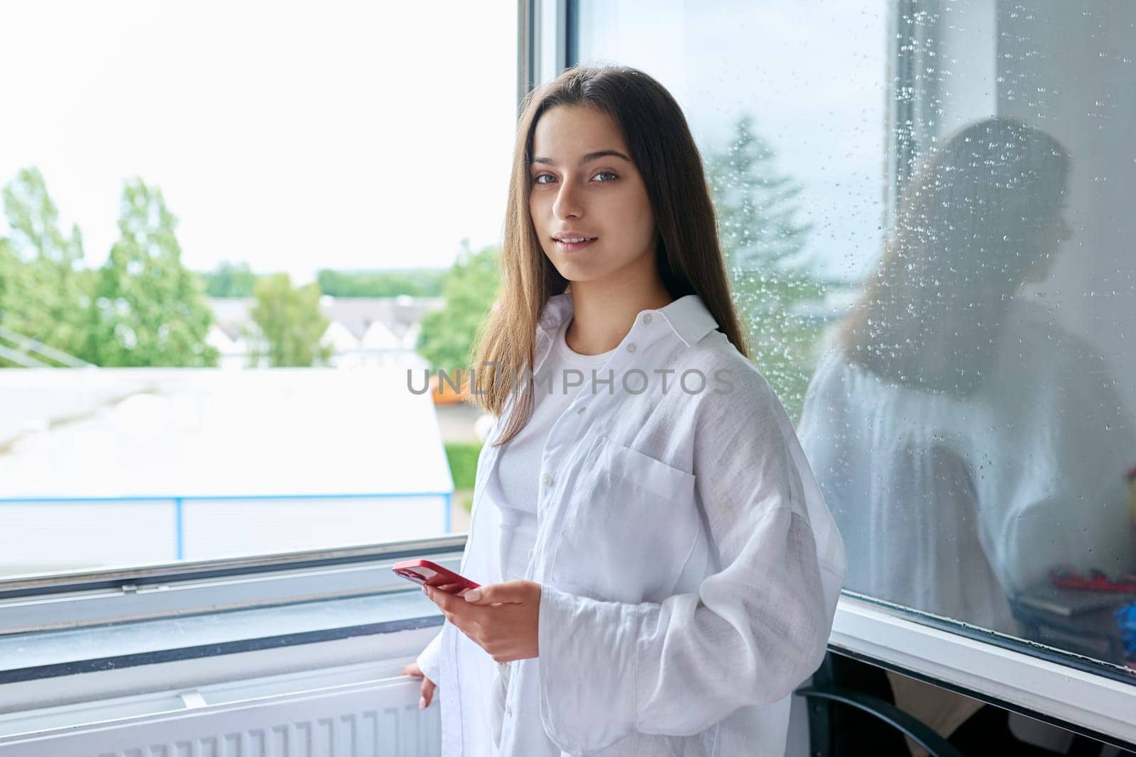 Portrait of a teen girl at home near window. Smiling female teenager 16, 17 years old looking at camera
