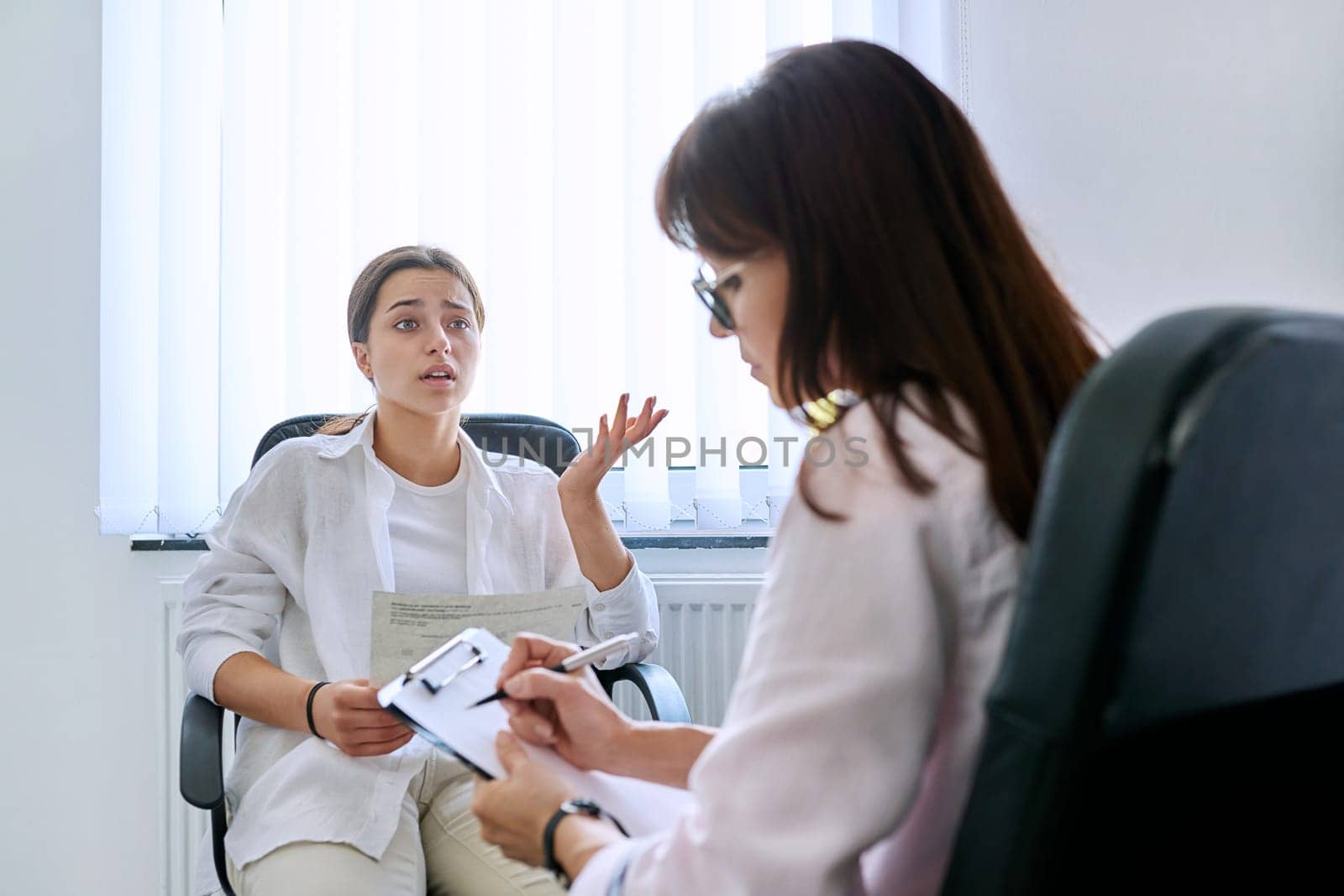 Female teenager at therapy meeting with professional psychologist terapist. Young sad serious girl showing formal mail letter. Psychology, psychotherapy, counseling, mental health of adolescents
