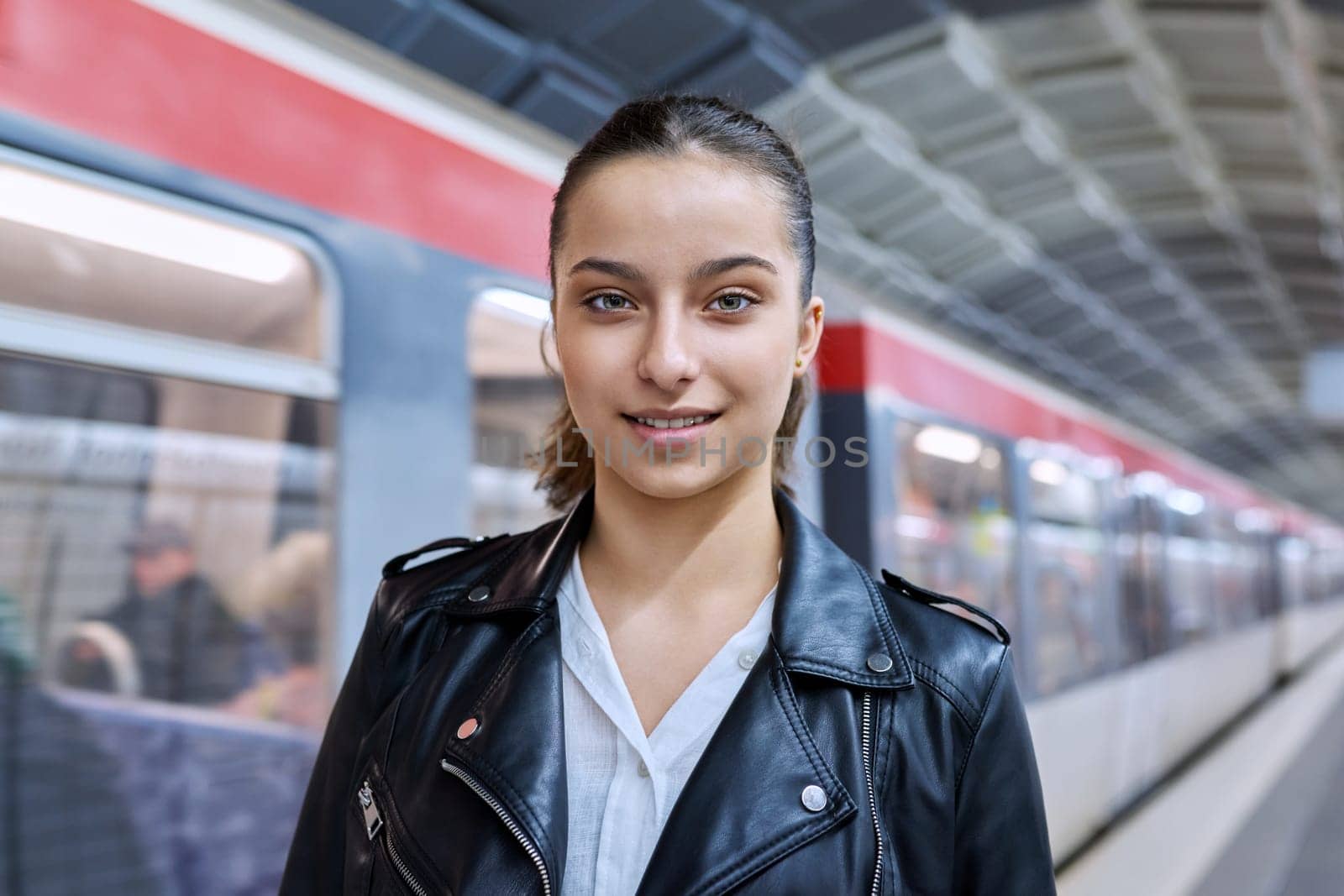 Teenage girl on platform of subway station, against backdrop of subway train, smiling beautiful female looking at camera. Student teenager youth, lifestyle in city, subway passengers, urban transport