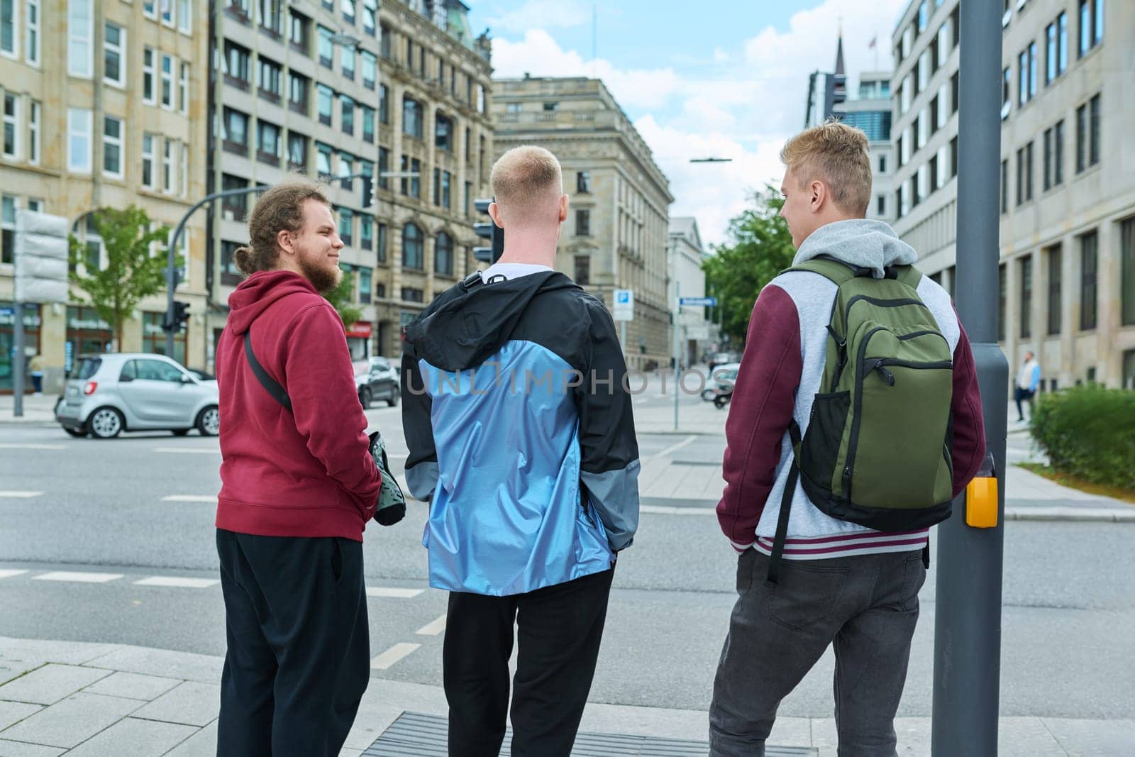 Back view of three friends of young teen boys college students on the city street. Teenagers at a crosswalk waiting for the green light of a traffic light