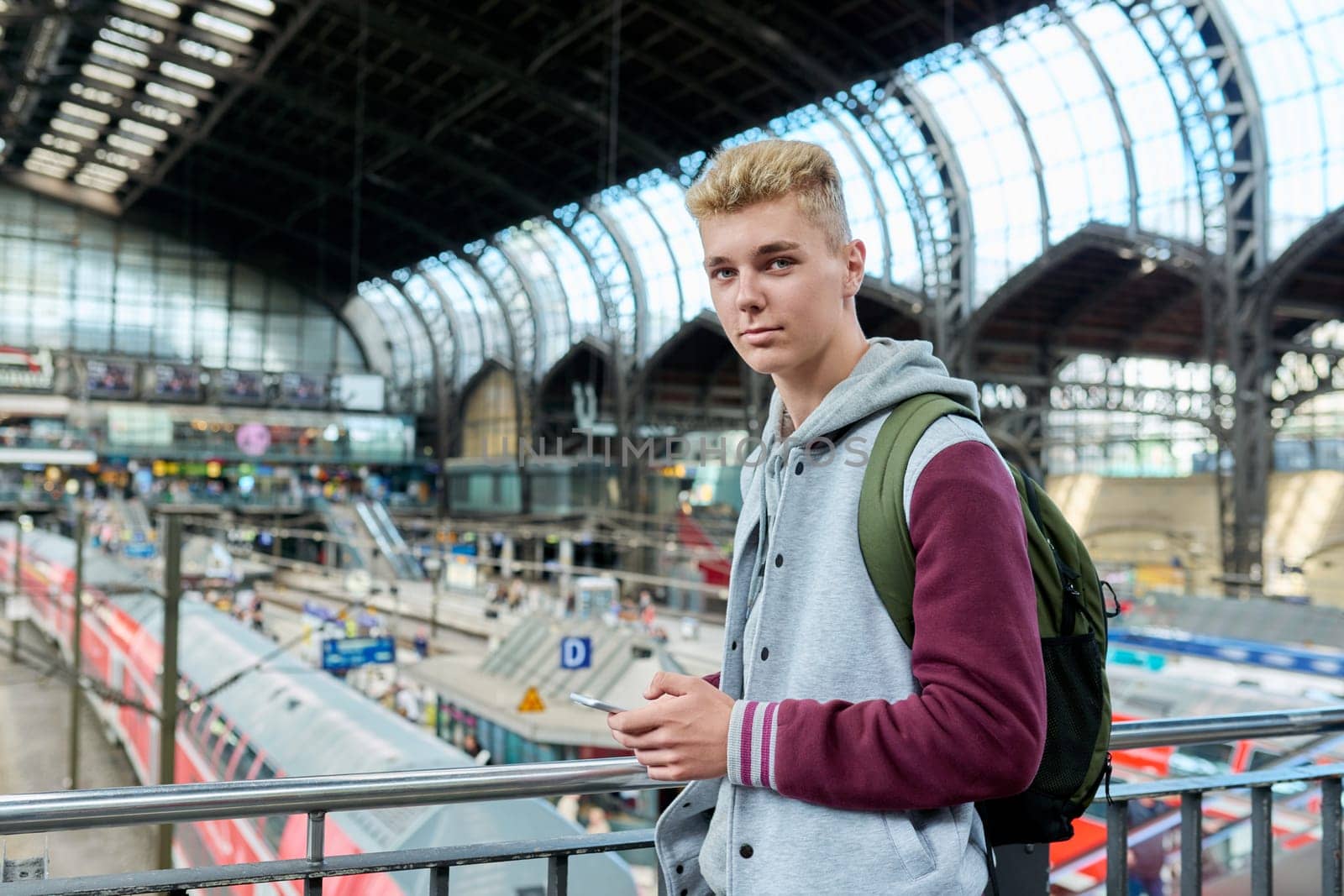 Portrait of smiling young guy with backpack smartphone at train station by VH-studio