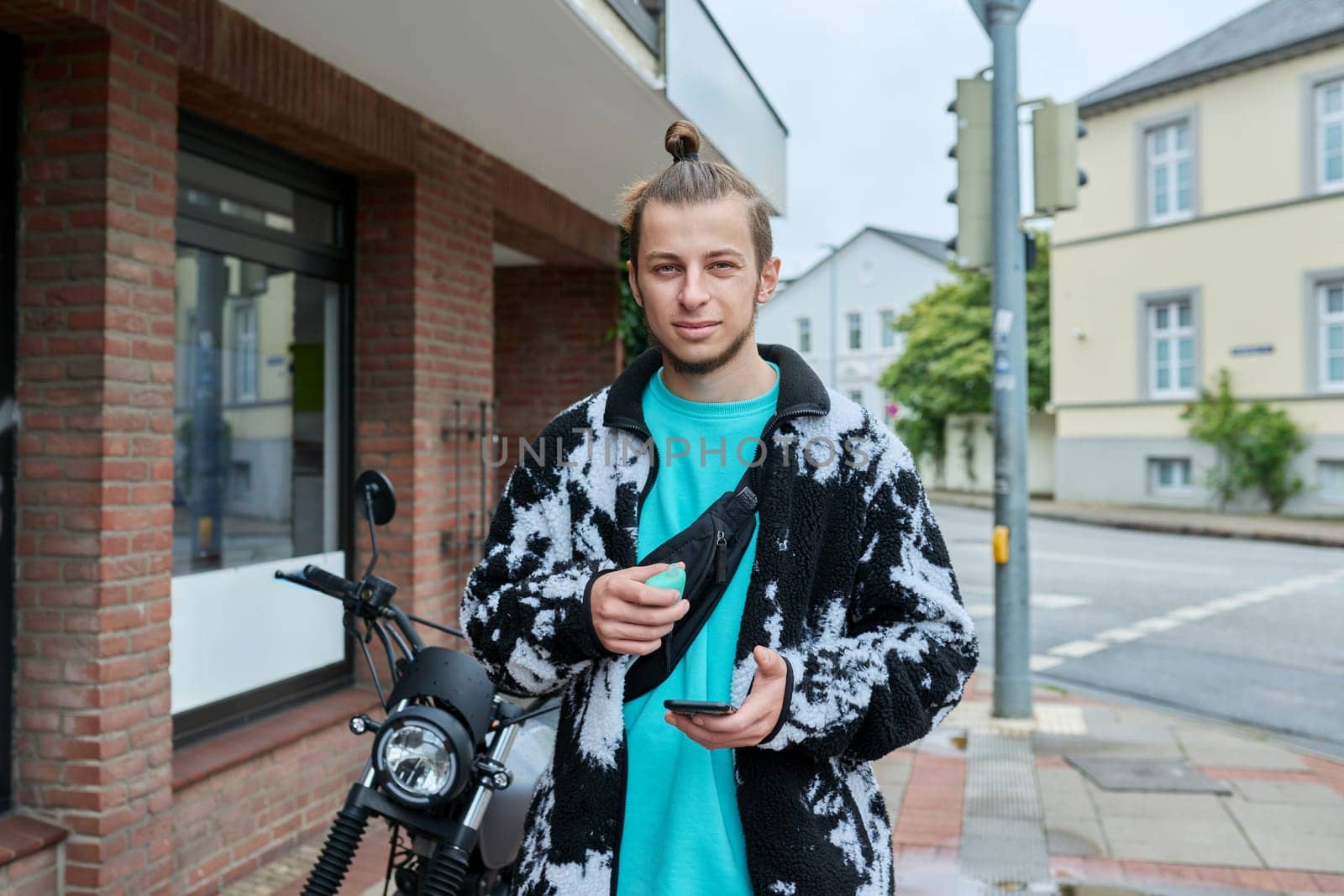 Young trendy male with phone and an electronic cigarette standing on motorcycle on city street. Youth, lifestyle concept