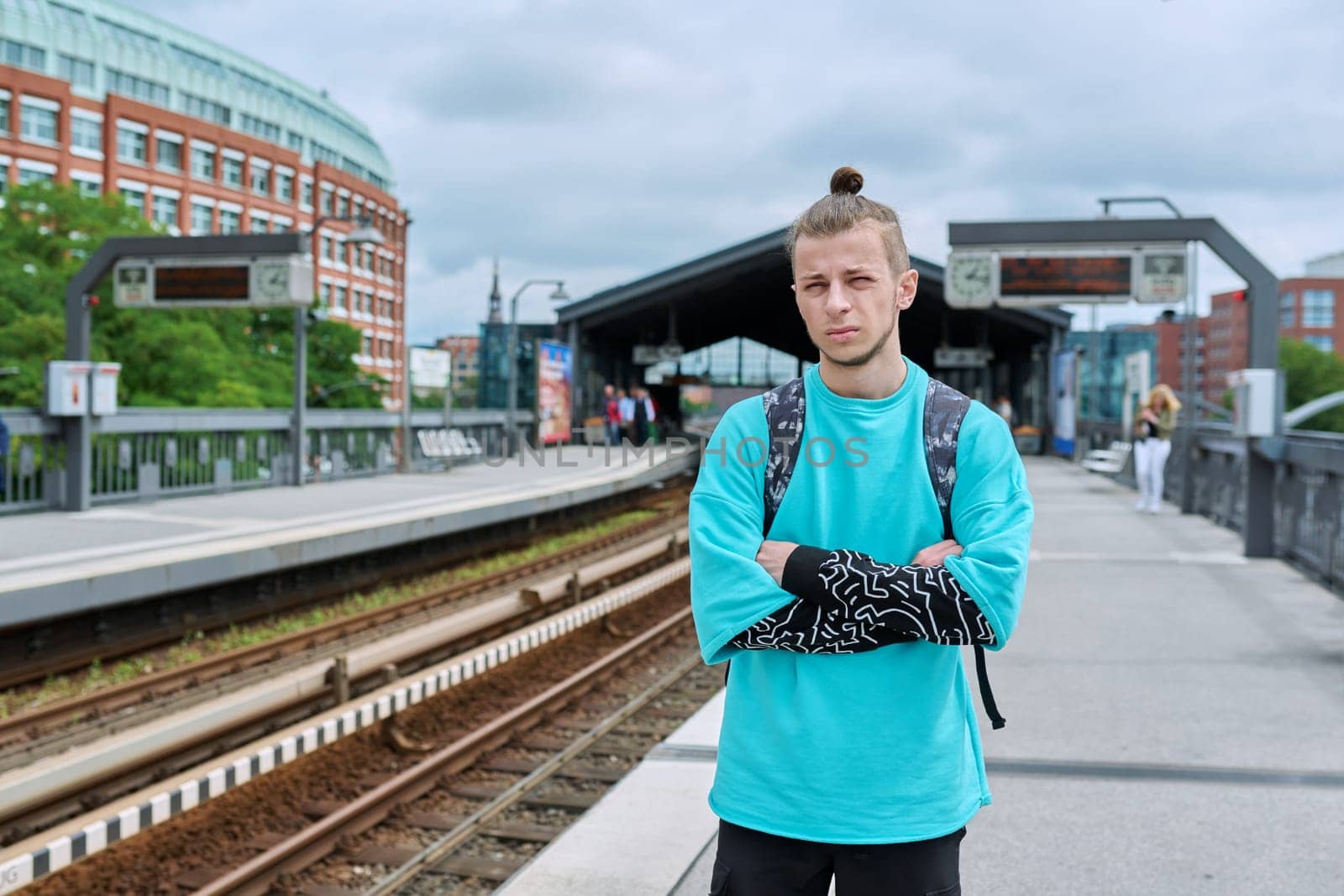 Young male waiting for electric train at city railway platform station. Guy serious student hipster 19, 20 years old with backpack. Urban transport, commuter train, passenger transportation, youth