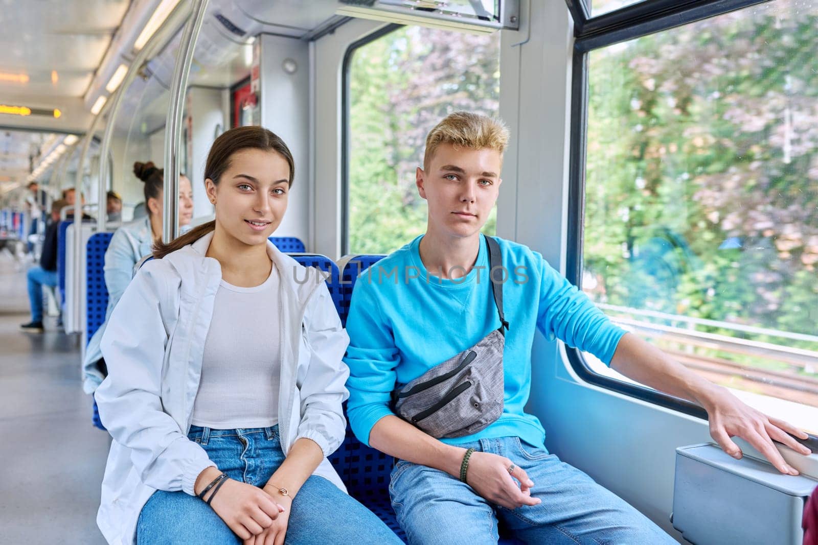 Teenage guy and girl commuter train passengers sitting together, smiling, looking at camera. Adolescence, youth, lifestyle, friendship, communication, rail transport, passenger traffic