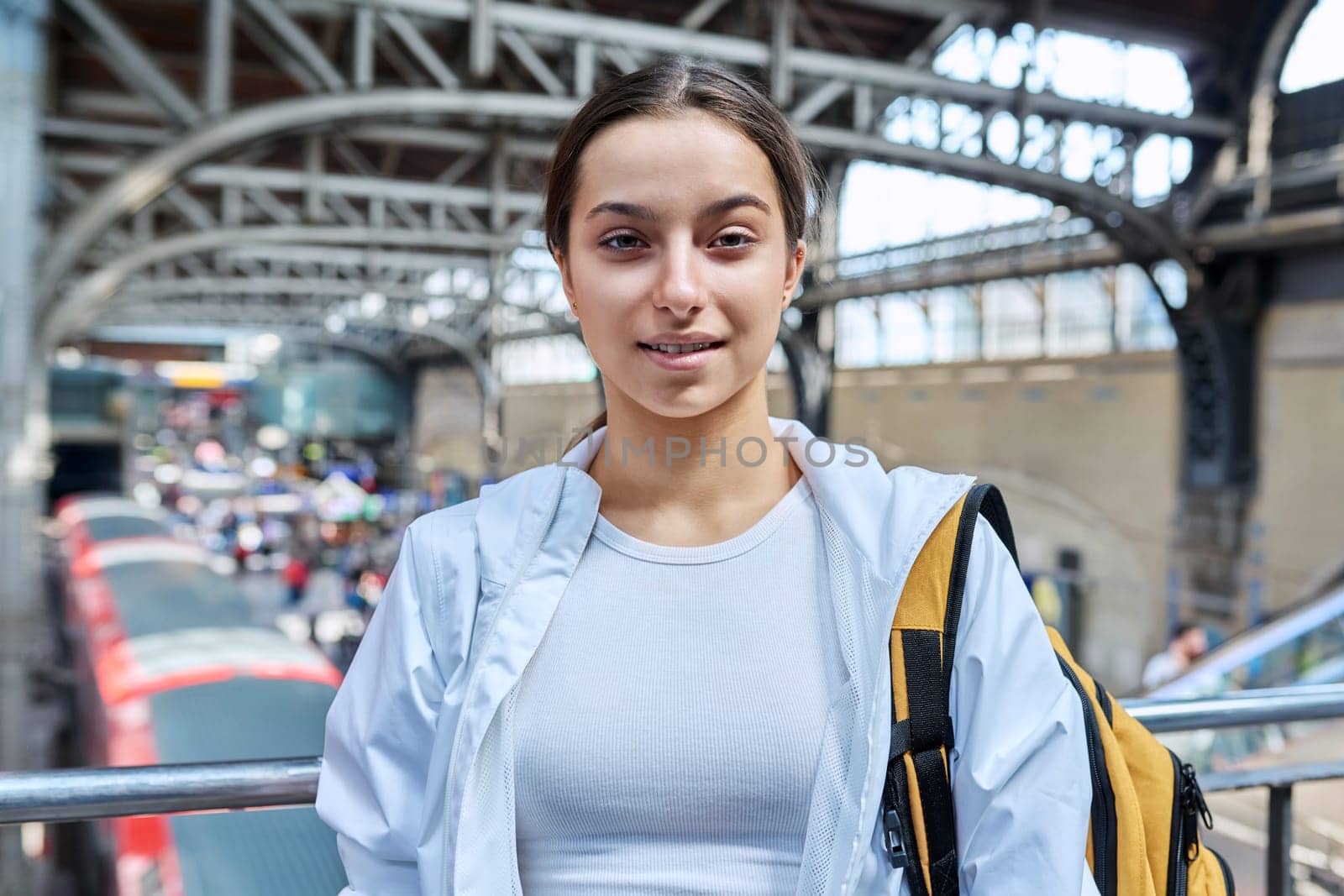 Teenage girl with backpack looking at camera at railway station. High school student, teenager girl passenger, suburban urban and intercity rail transport