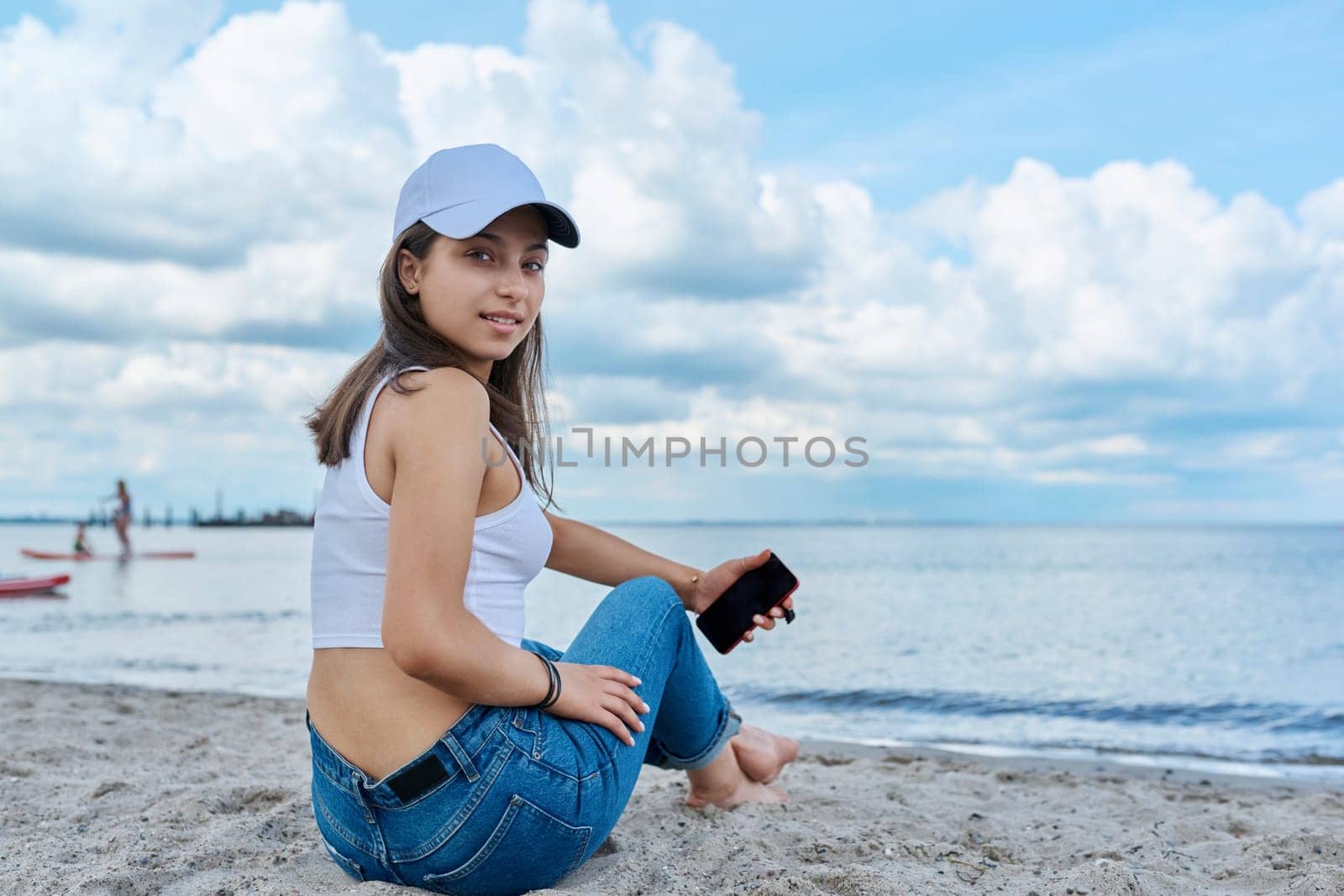 Young female in jeans cap sitting on the beach, using smartphone, back view. Recreation, relaxation, nature, tourism, youth concept