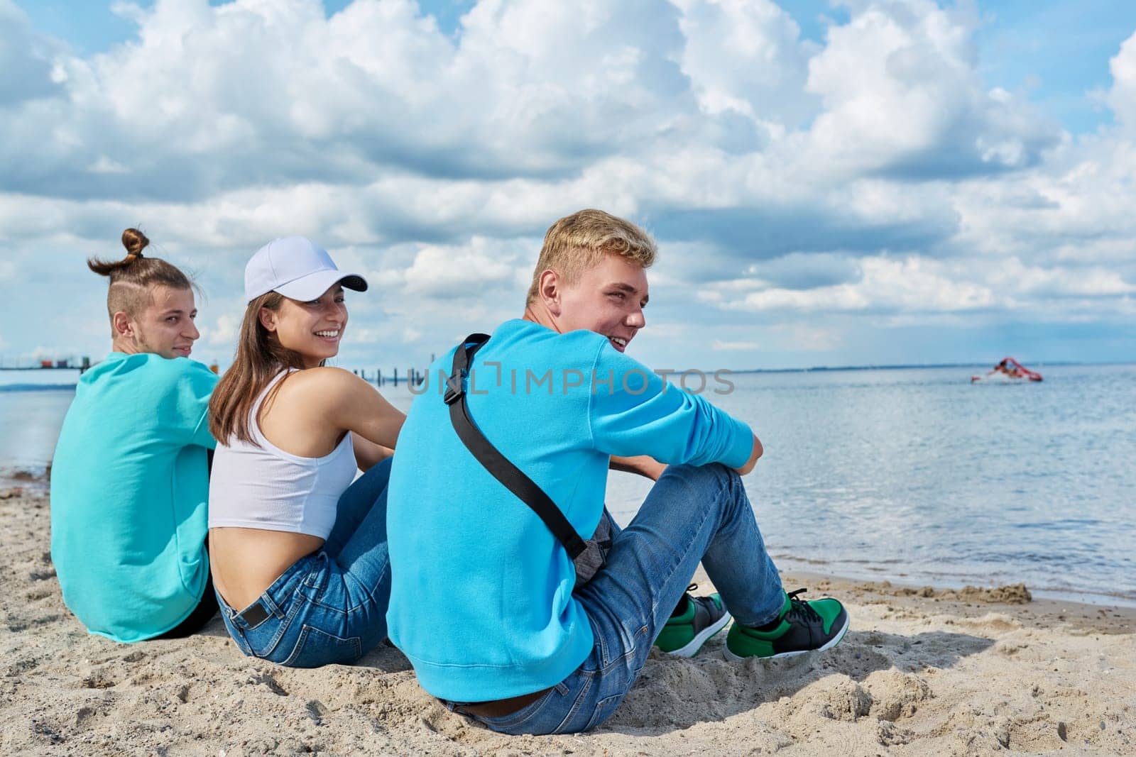 Back view of three teenage friends sitting at beach, looking at sea by VH-studio