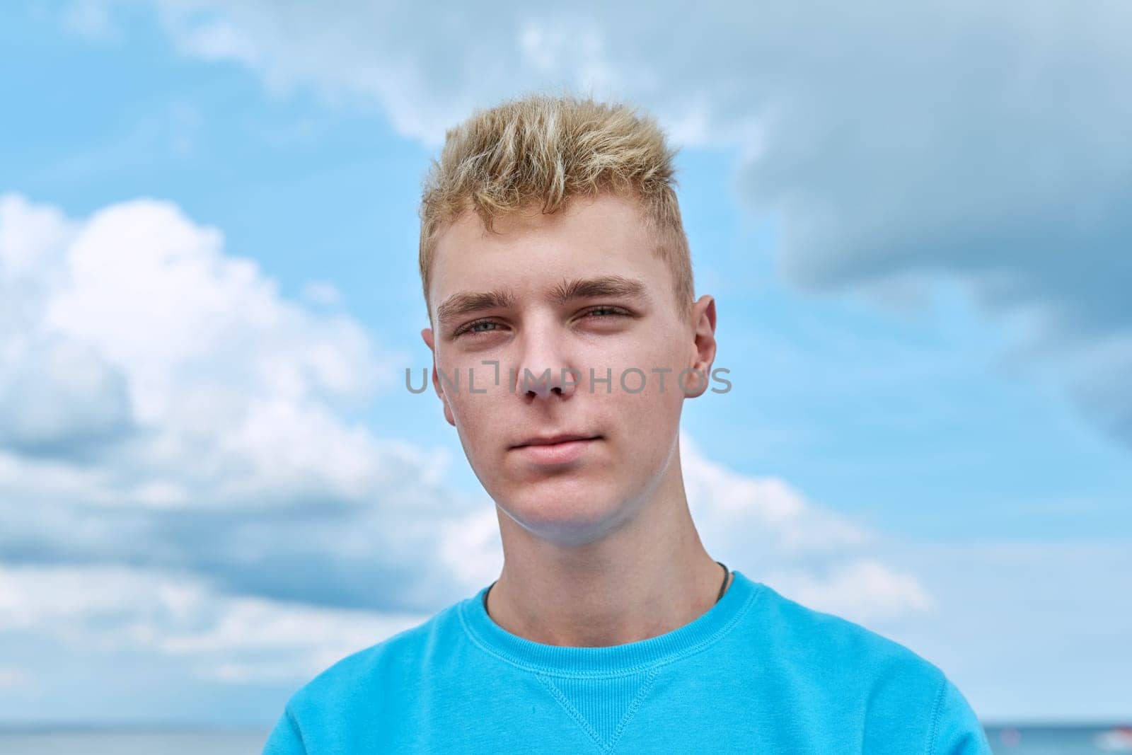 Headshot portrait of handsome smiling teenage guy, face of positive young male against blue sky in clouds. Youth, 18, 19, 20 years of age, lifestyle, enjoy fun, holiday vacation, outdoor