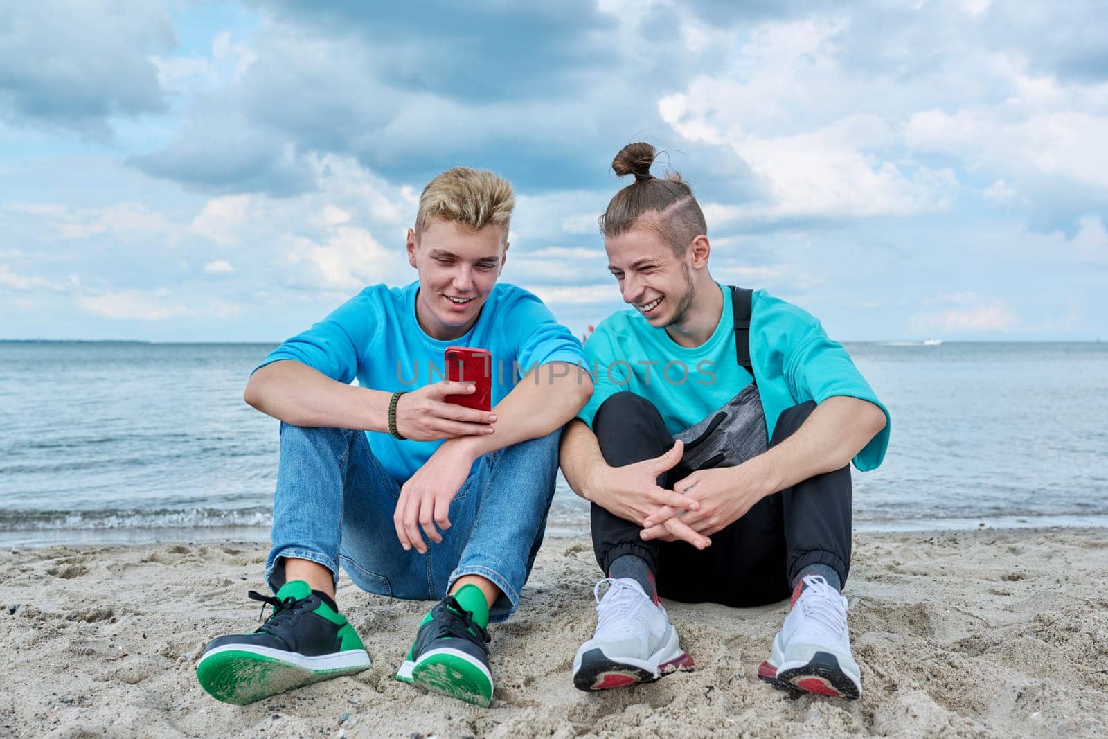 Young teenage male friends sitting on sand at beach, talking, laughing, looking in smartphone. Youth, summer vacation, communication, relaxing, leisure, friendship, fun, lifestyle holiday concept