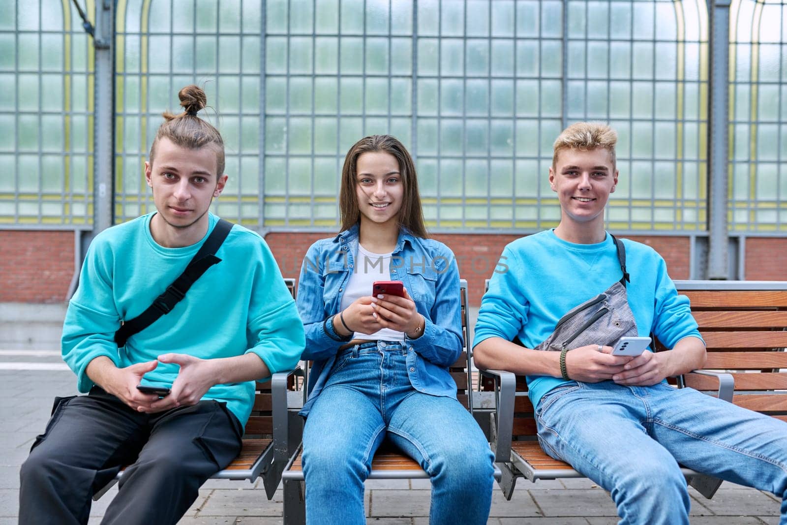 Group of teenage friends using smartphones sitting on chairs on train station platform, looking at camera. Youth, adolescence, digital technology, mobile applications, communication learning networks