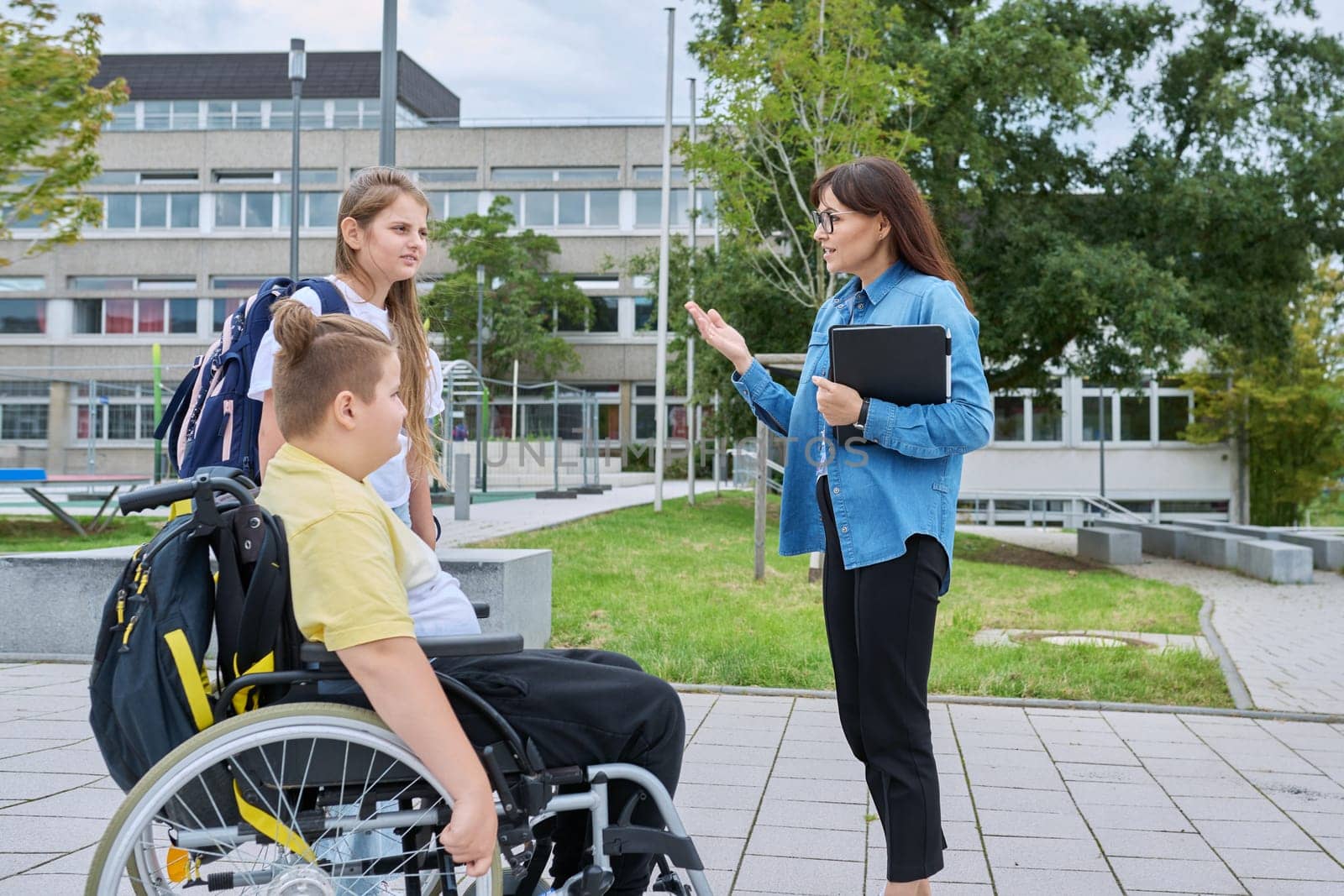 Female teacher and children, boy in wheelchair and girl talking outdoor by VH-studio