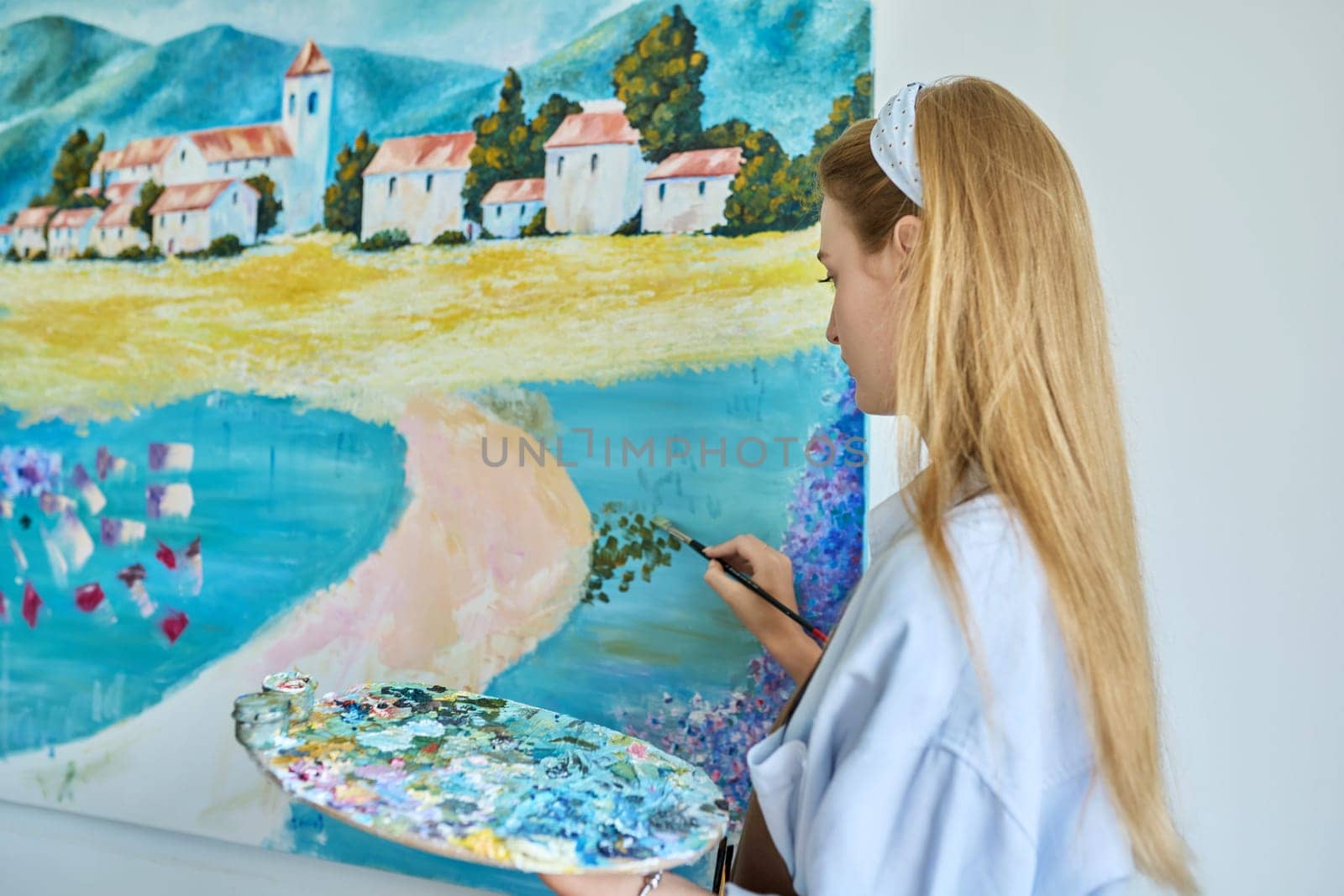 Young woman artist with acrylic paint palette with brushes painting picture on canvas on wall. Creativity, hobby, art therapy, leisure, paintings for sale, youth concept