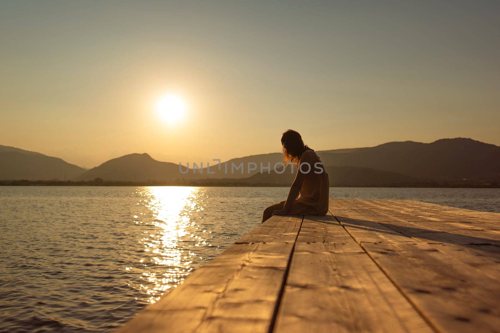 A thoughtful young Caucasian woman sits alone on a lake pier, gazing at the water during a sunset, in a vintage orange ambiance.