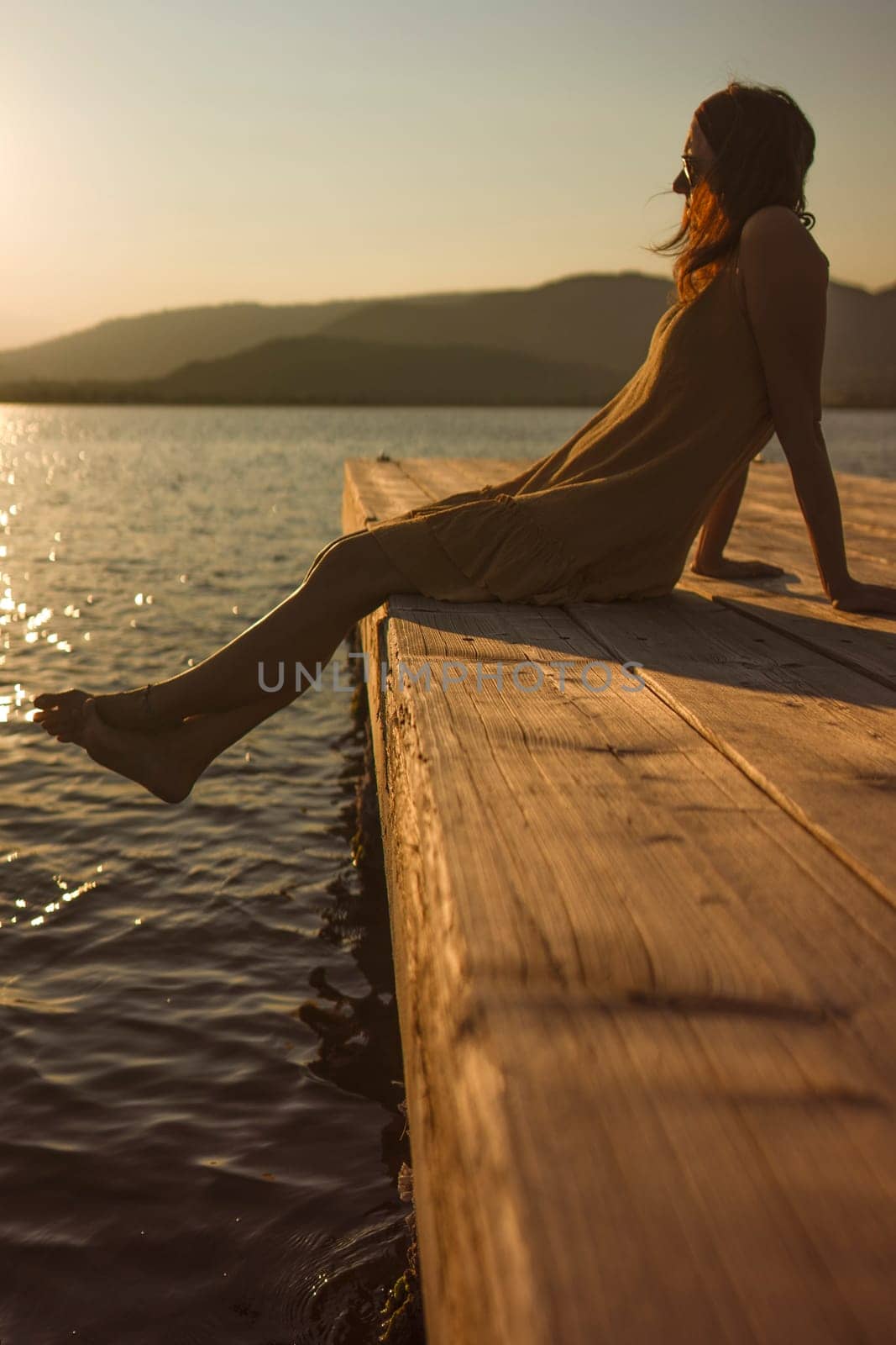 A contemplative young Caucasian woman sits alone on a lakeside pier, gazing at the water at sunset or dawn, in orange and vintage atmosphere. by robbyfontanesi