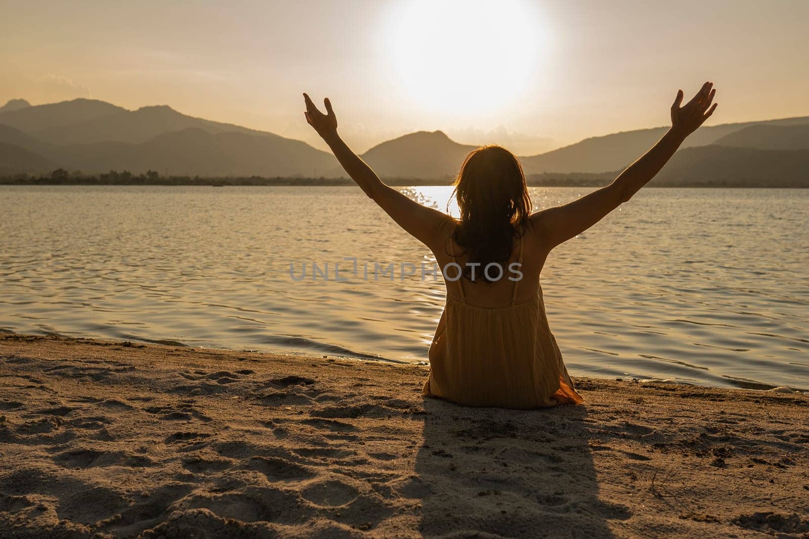Evocative shot: woman in contemplative pose sitting on the sand with arms wide, facing the sea at sunrise or sunset on a beach. Connect with nature.