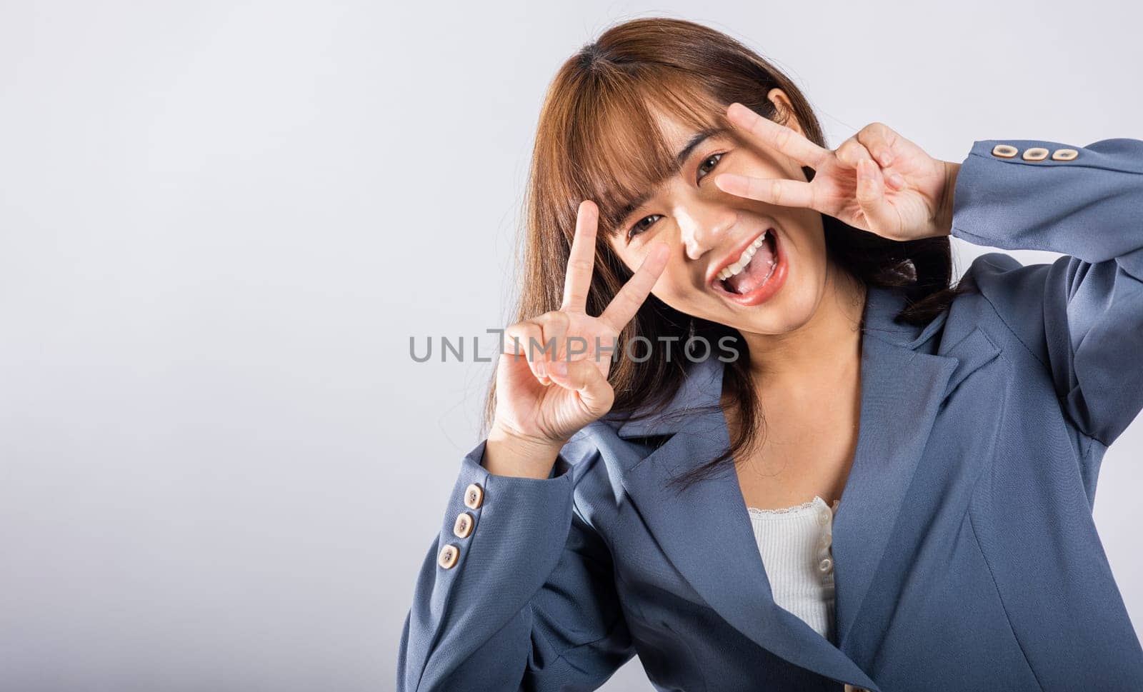 An Asian woman showcases her optimism and peace by gesturing victory and peace signs near her eye in a studio shot with a white background. Her joyful smile speaks of success.