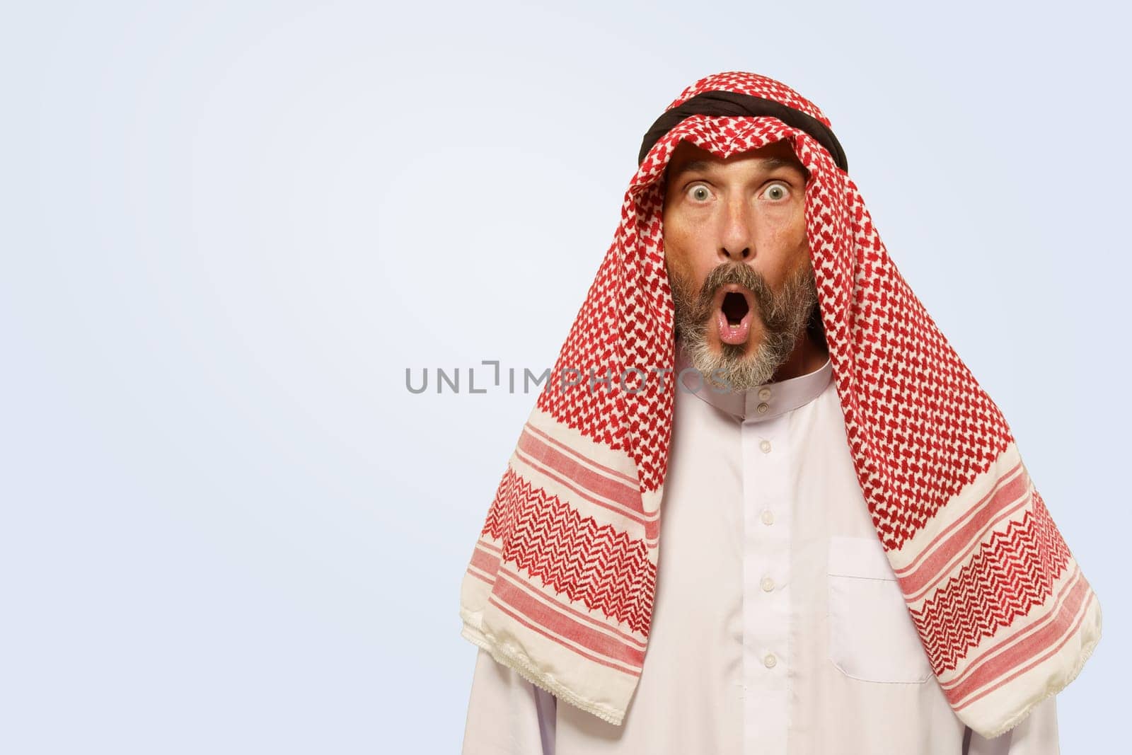 Arabian man, in keffiyeh, displays expression with shock, surprise and frustration, reflected by opened mouth. He isolated on gentle light blue background, illustrating cultural identity of ethnic group. High quality photo