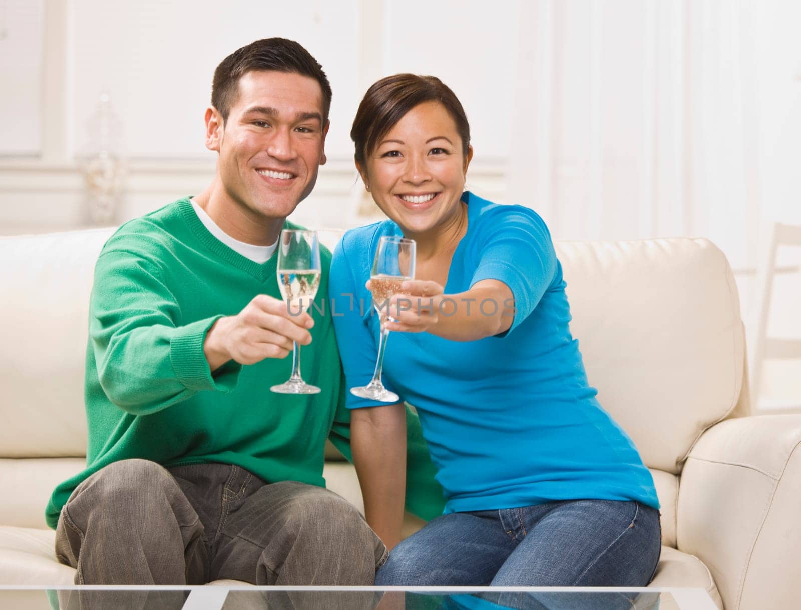An attractive young asian couple toasting champagne. They are seated on a couch together and are smiling at the camera. Horizontally framed shot.