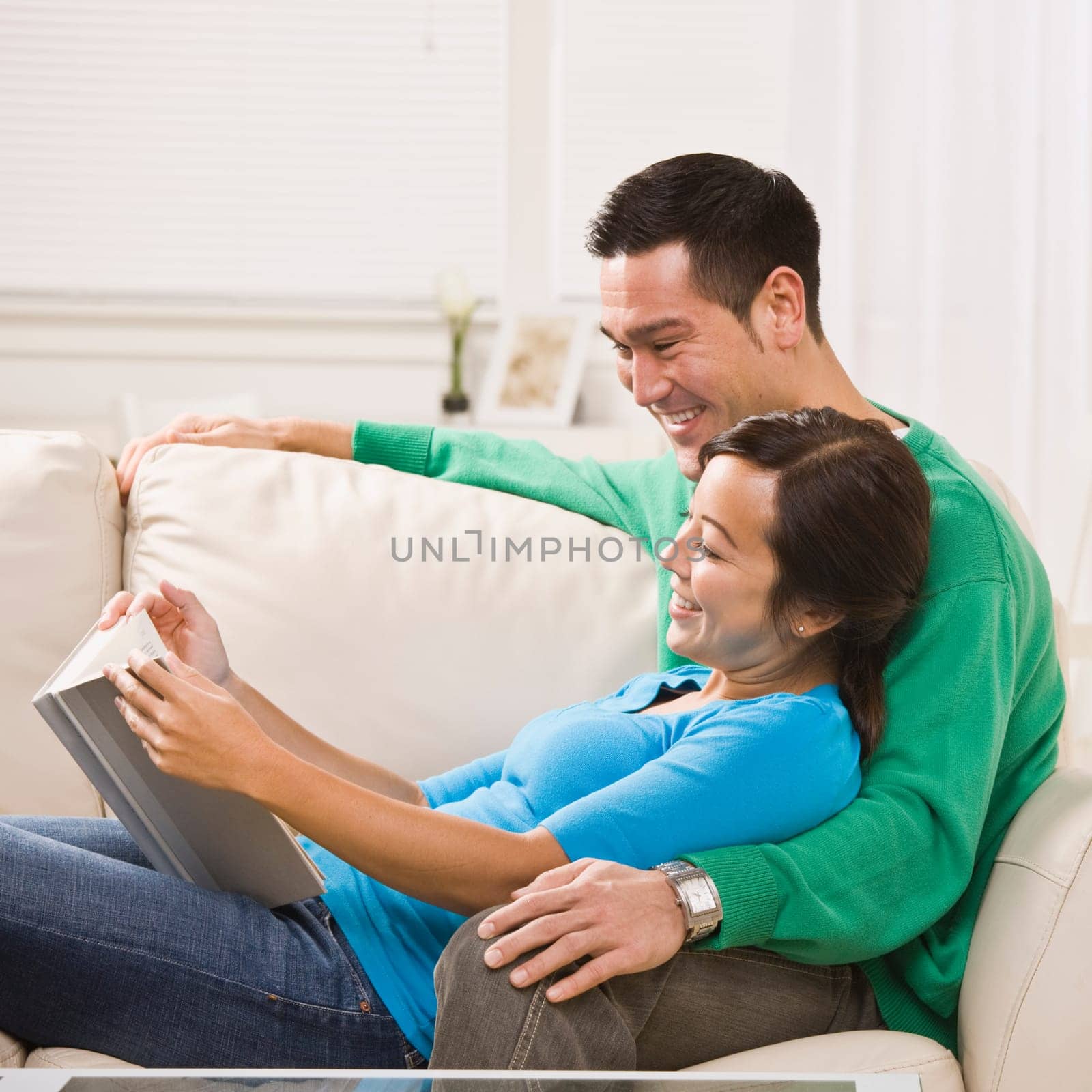 A young couple is seated together on their living room couch. They are reading a book together, smiling, and looking away from the camera. Square framed shot.