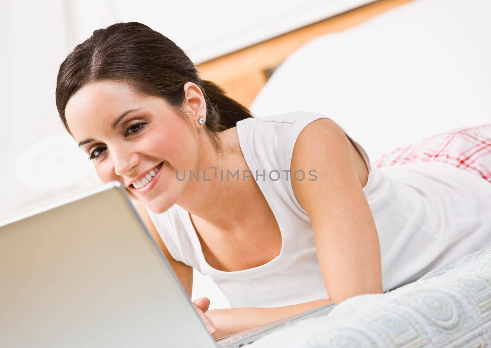 A beautiful young woman using a laptop in bed. She is smiling at the laptop screen. Horizontally framed shot.