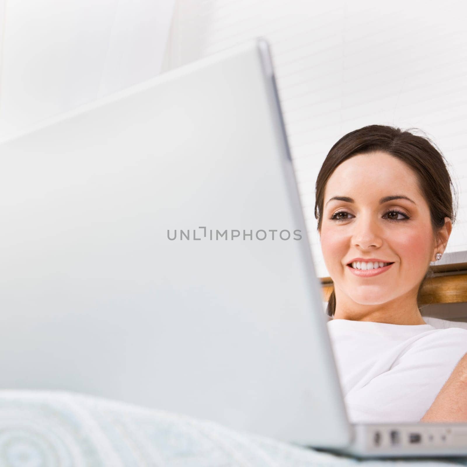 A beautiful young woman is sitting in a room and working on a laptop. She is smiling and is looking at the screen. Square framed shot.