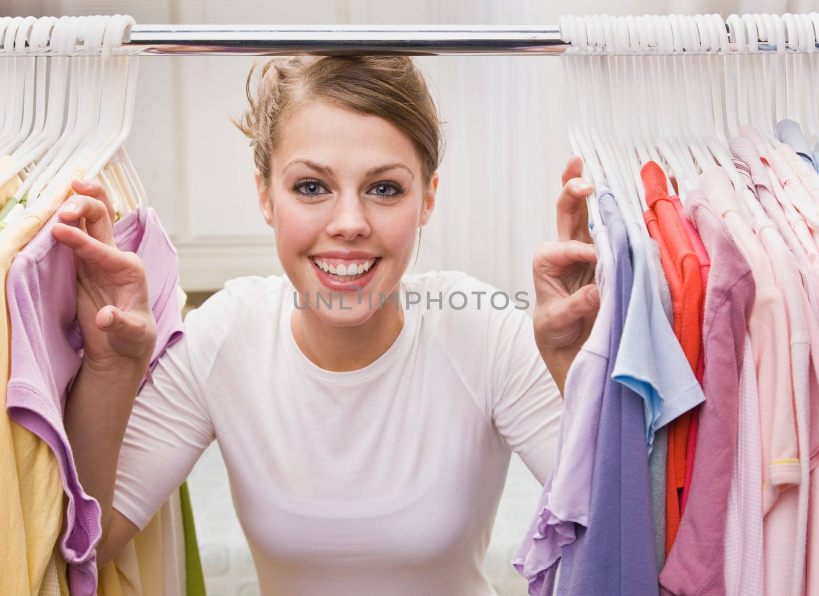 A young, attractive woman is standing in a closet and looking through clothes. She is smiling at the camera. Horizontally framed shot.