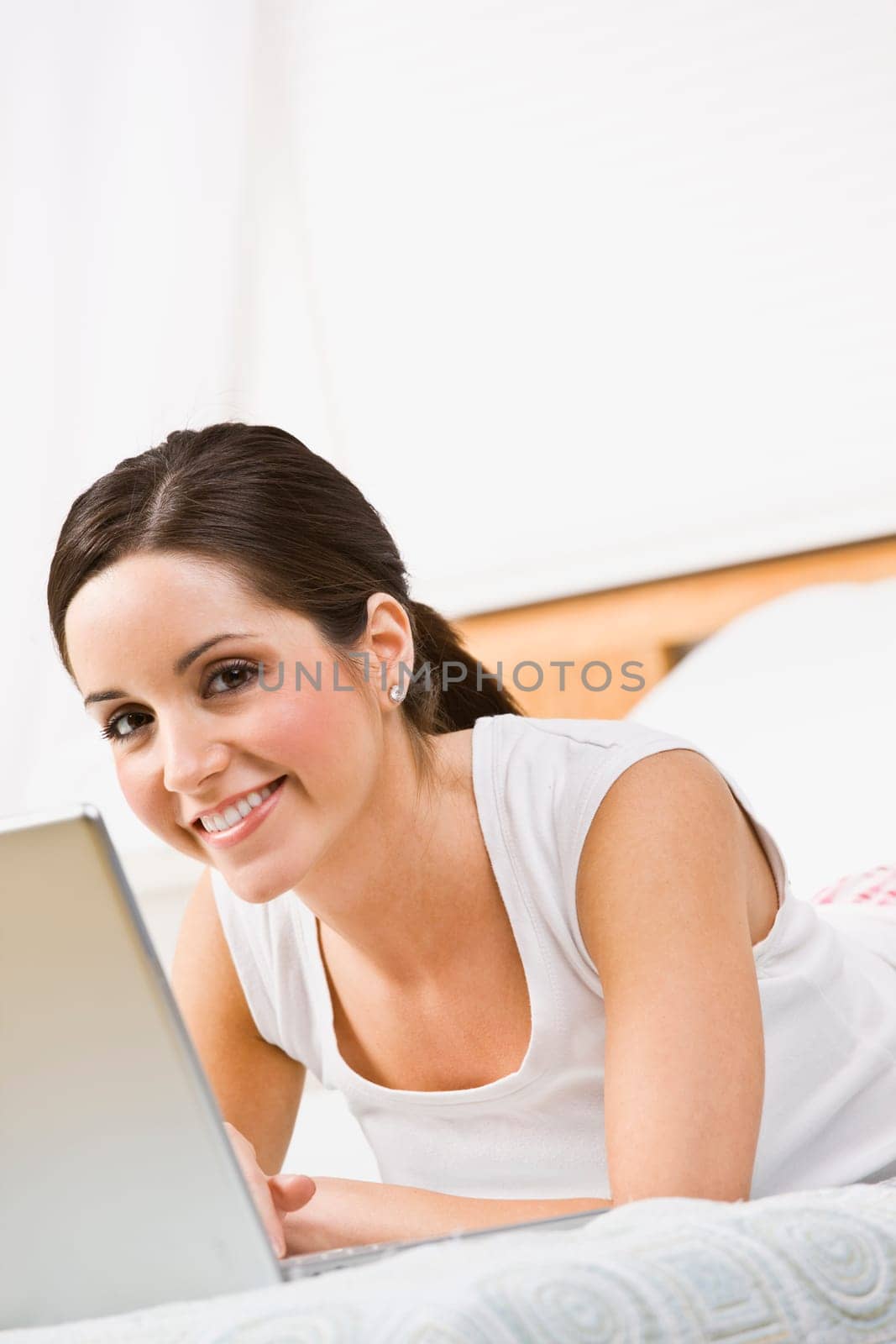 A beautiful young woman using a laptop in bed. She is smiling at the camera. Vertically framed shot.