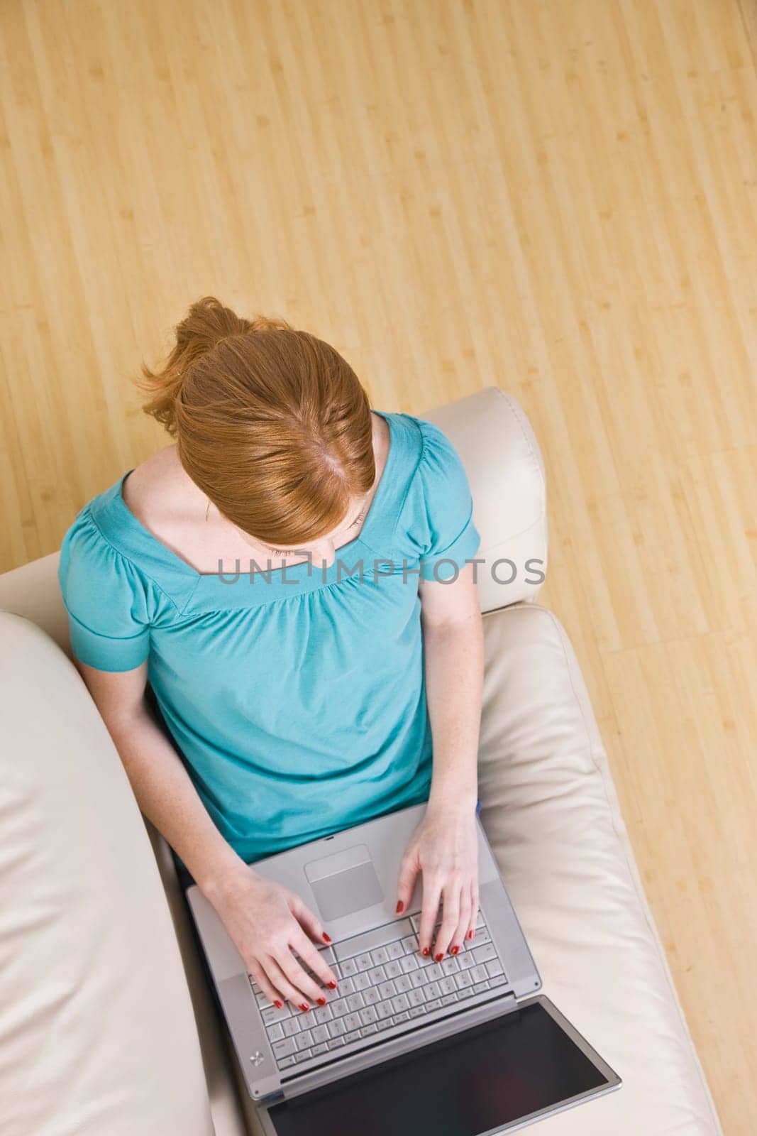 The above view of a young woman seated on a couch and working on a laptop. She has red hair. Vertically framed shot.