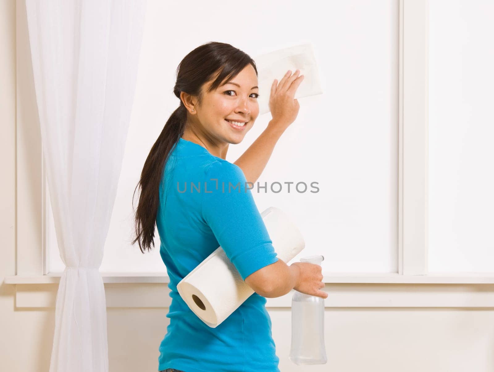 An attractive asian young female washing a window. She is holding a roll of paper towels and has her head turned to smile toward the camera. Horizontally framed photo.