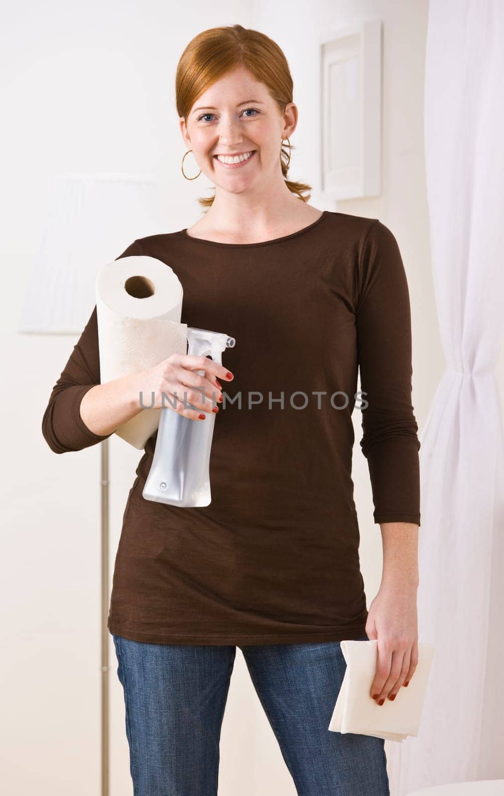 An attractive young woman holding cleaning supplies. She is smiling directly at the camera. Vertically framed photo.