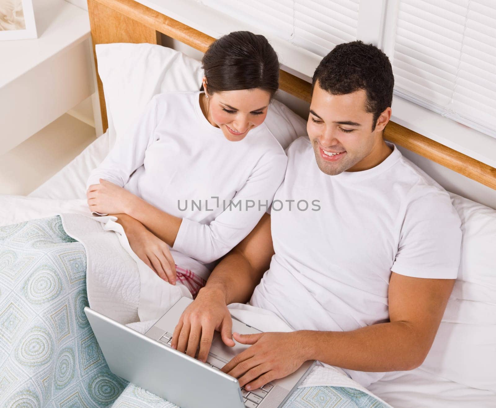 An attractive young couple lying in bed together and viewing a laptop screen. They are both smiling. Horizontally framed shot.