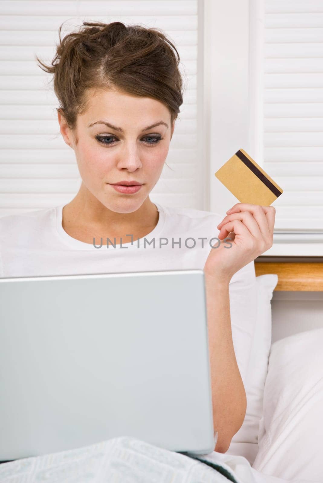 An attractive young woman holding a credit card and peering at a laptop screen. She is sitting in bed and has a contemplative expression. Vertically framed shot.