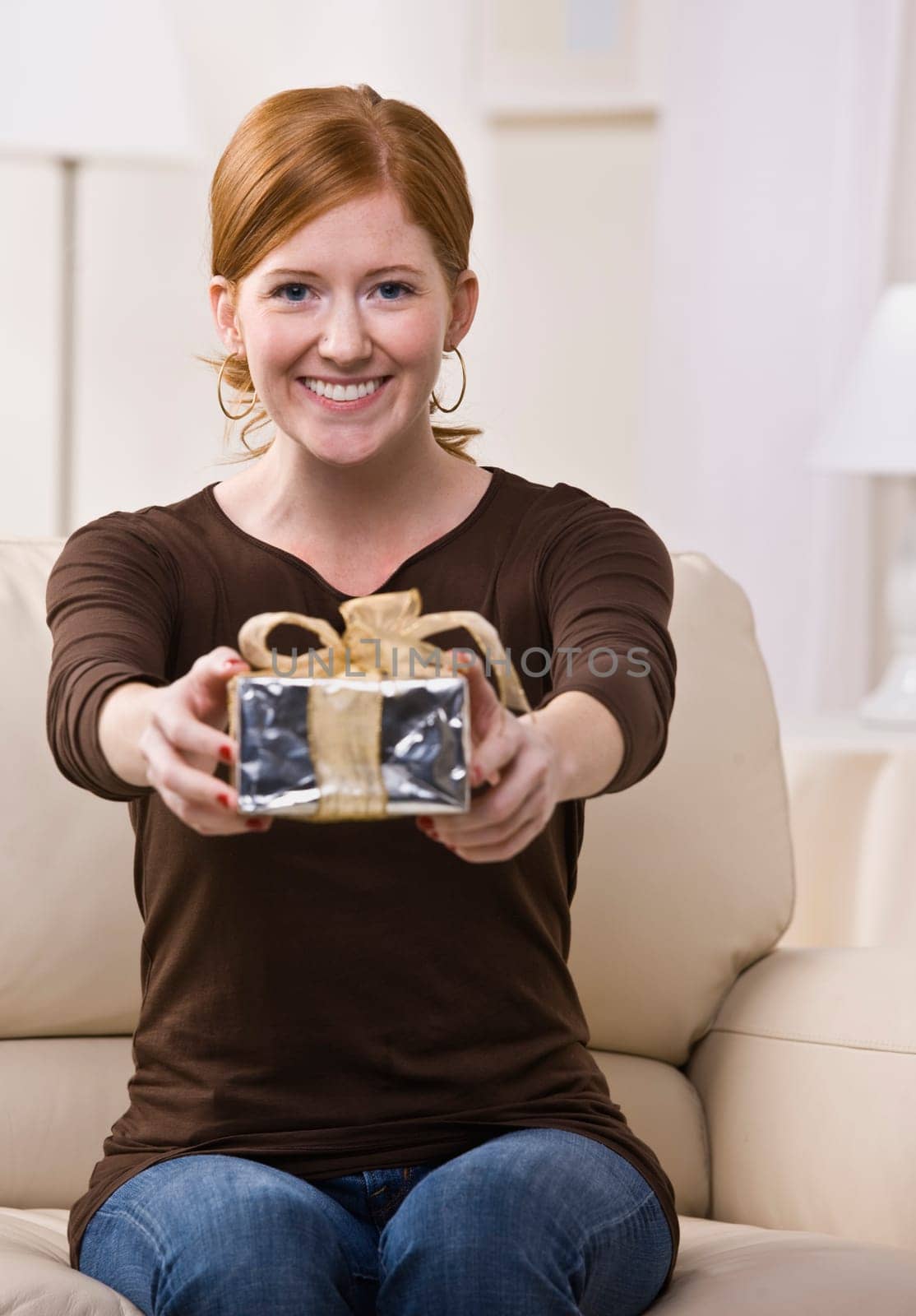 An attractive young female sitting on a couch and presenting a gift with outstretched arms. She is looking directly at the camera and is smiling. Vertically framed photo.