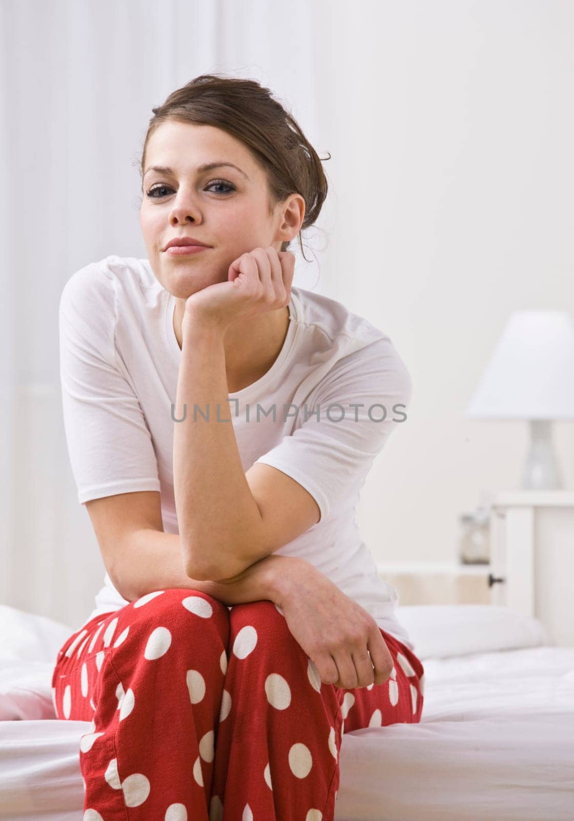 An attractive young woman sitting on the edge of a bed in her pajamas. She is resting her chin on her hand and is smiling slightly toward the camera. Vertically framed photo.