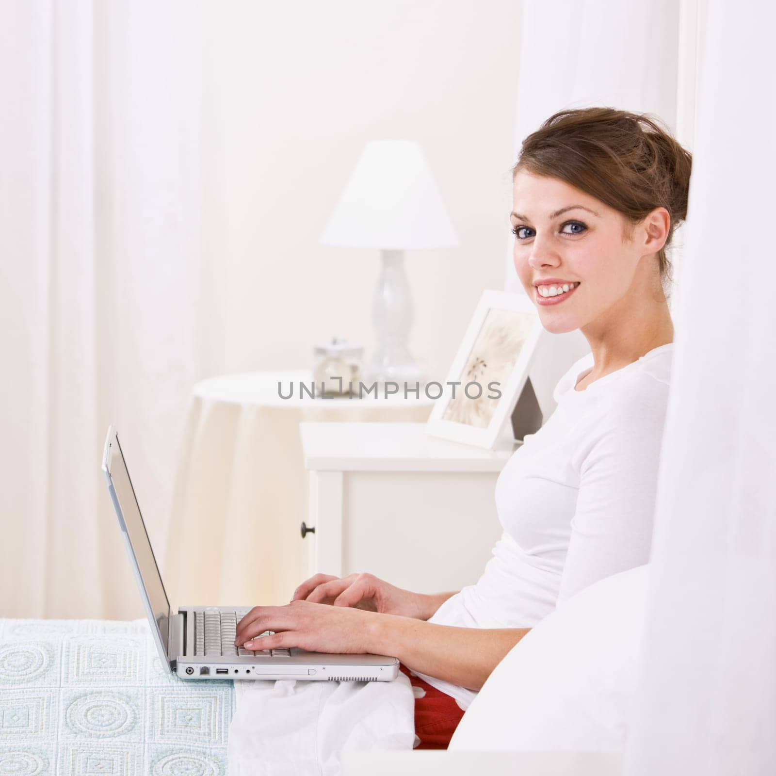An attractive young female lying in bed and using a laptop. She is smiling at the camera. Square framed photo.