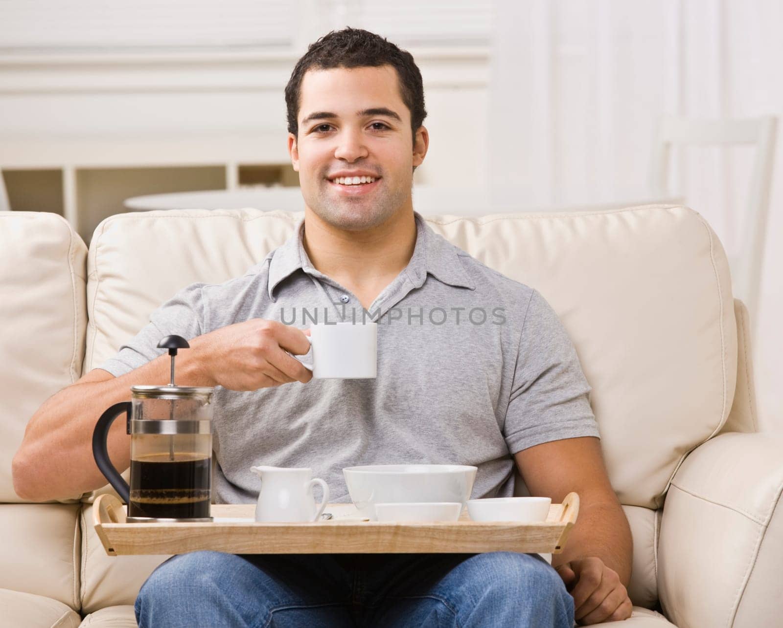 An attractive young man eating from a breakfast tray. He has a coffee cup raised, and is smiling directly at the camera. Horizontally framed photo.