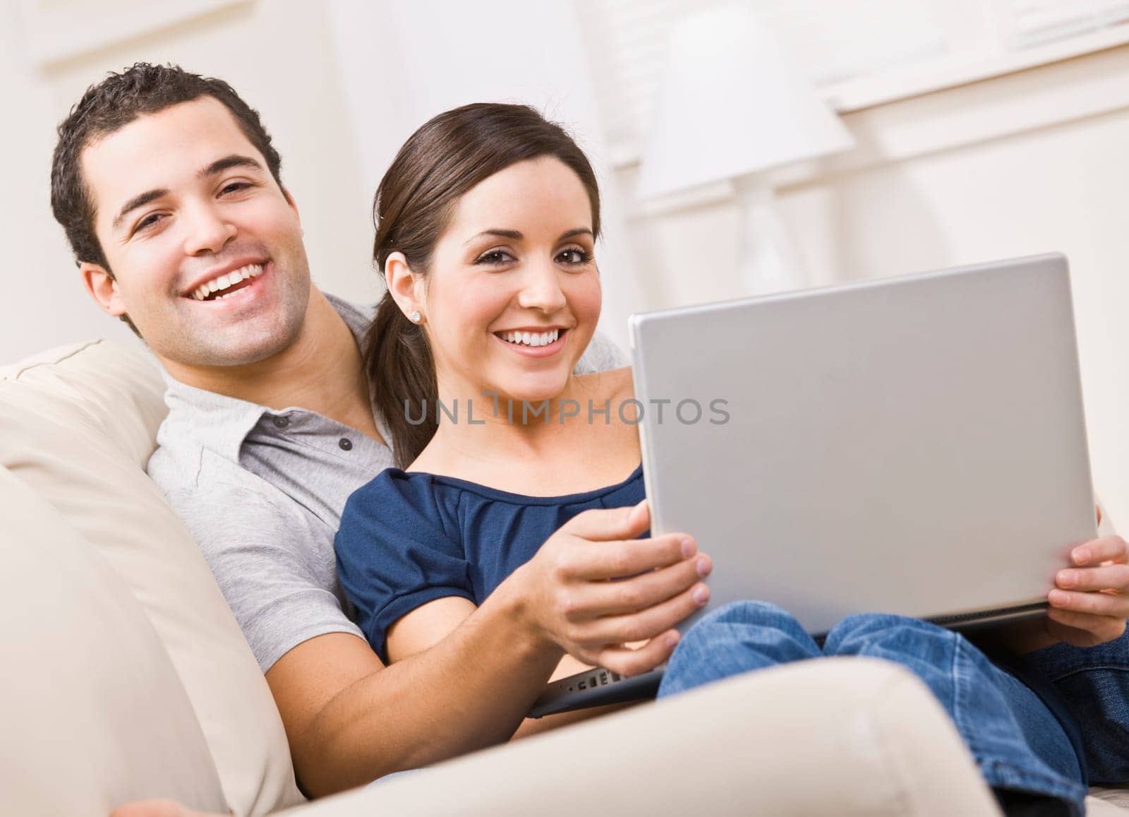 An attractive young couple sitting on a couch together and holding a laptop. They are smiling directly at the camera. Horizontally framed shot.