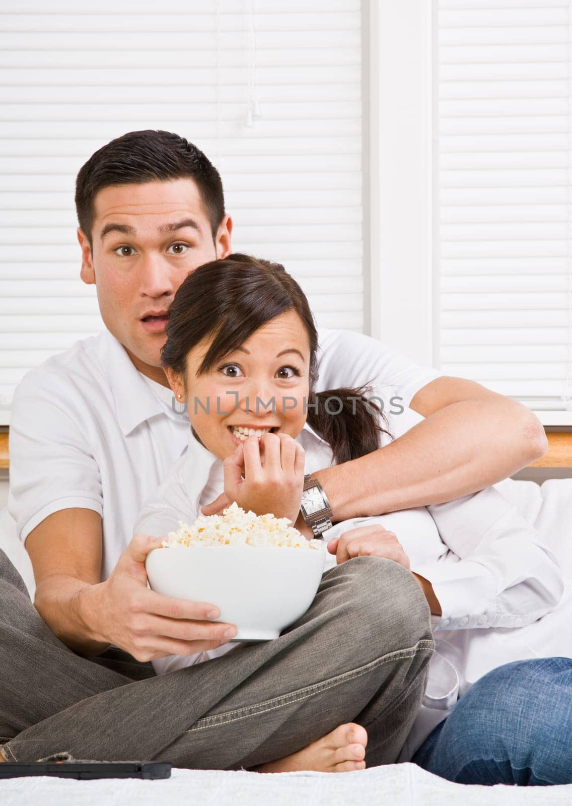 A young, attractive couple is sitting together in bed and watching TV. They look shocked or scared, and are looking at the camera. Horizontally framed shot.